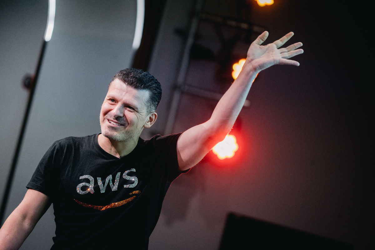 Smiling man wearing a black AWS t-shirt waving with his right hand on stage with bright red lights in the background.