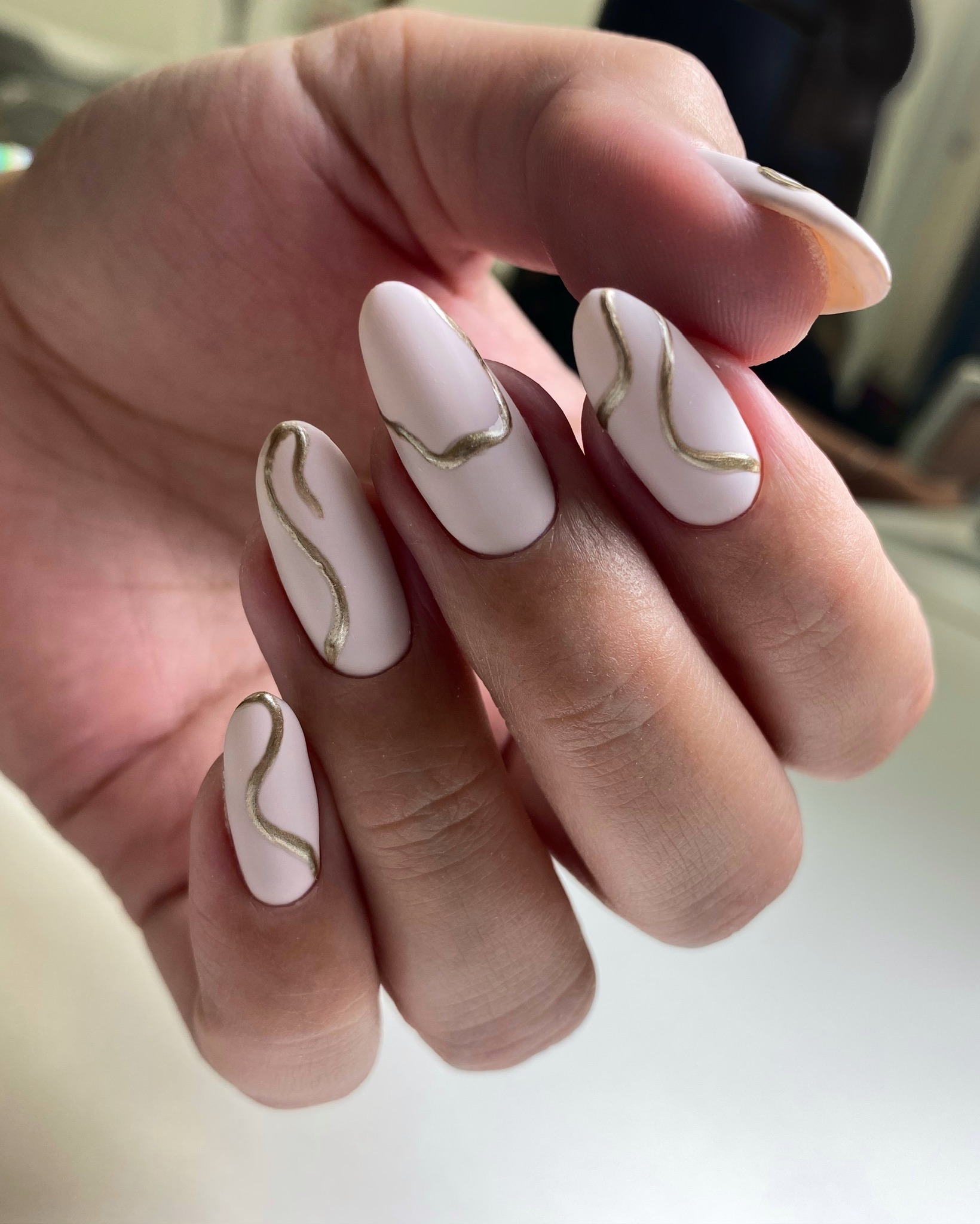 Hand with pale pink matte nails featuring thin gold wavy line nail art on each finger.