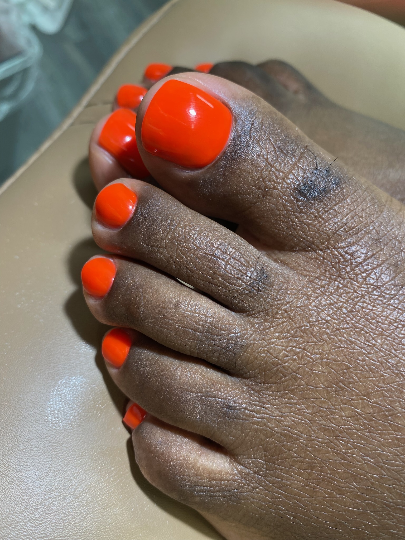 Close-up of dark-skinned toes with bright orange nail polish on beige surface.