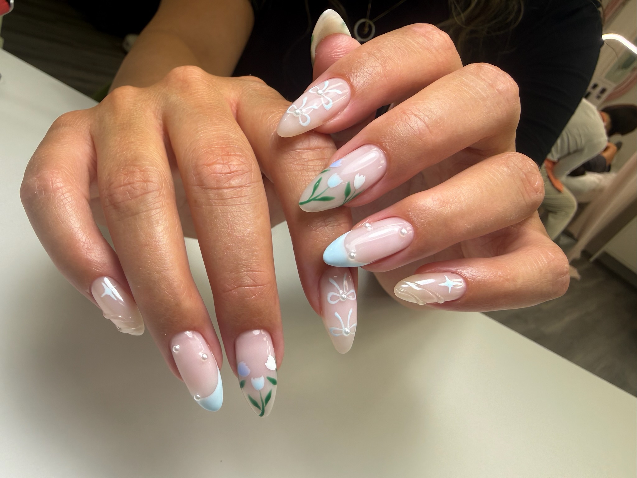 Close-up of hands with almond-shaped nails featuring pastel blue tips, white bows, flowers, and pearls.