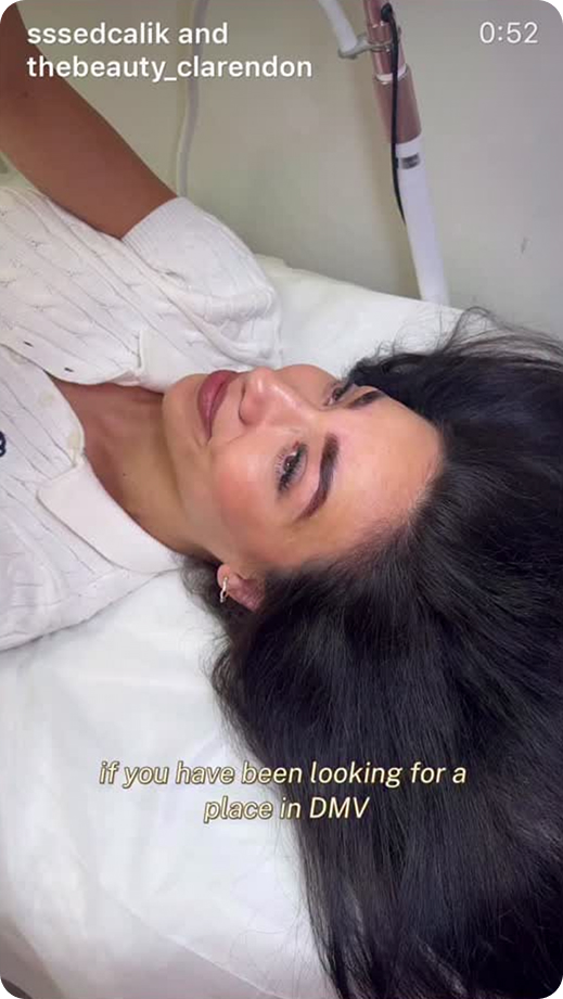 Woman lying on white bed with long dark hair, smiling slightly, wearing a white top.