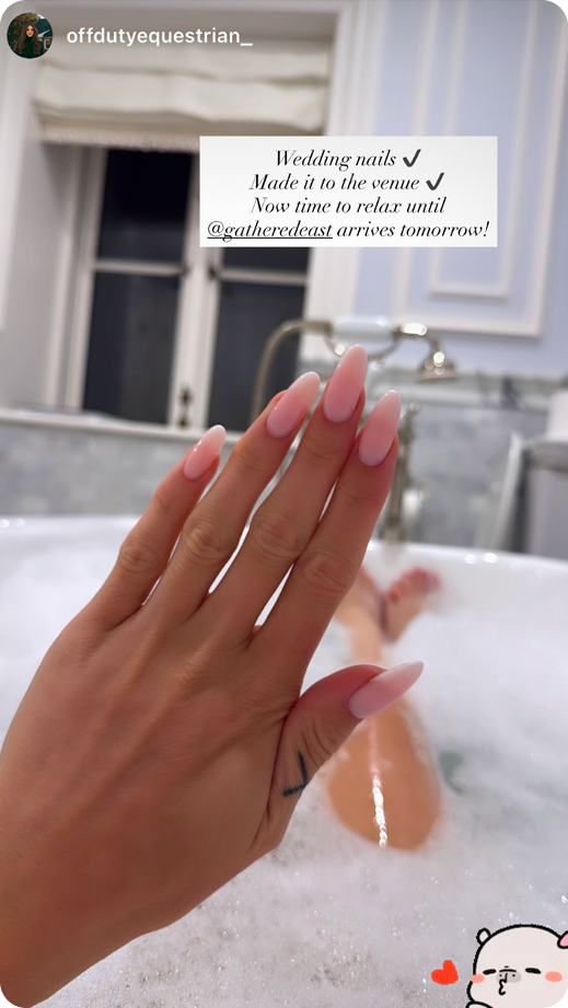 Hand with pink long matte nails over a bubble bath with feet visible in the tub.