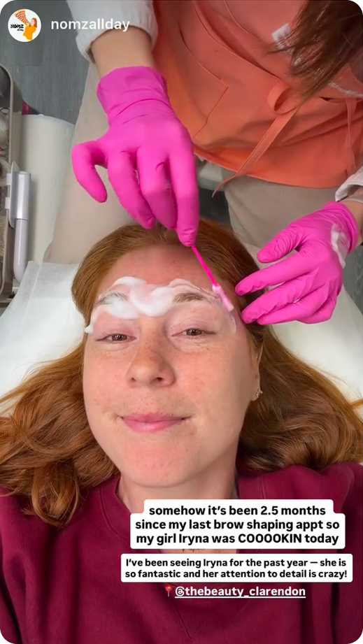 Woman lying down while a beautician in pink gloves shapes her eyebrows with a cotton swab.