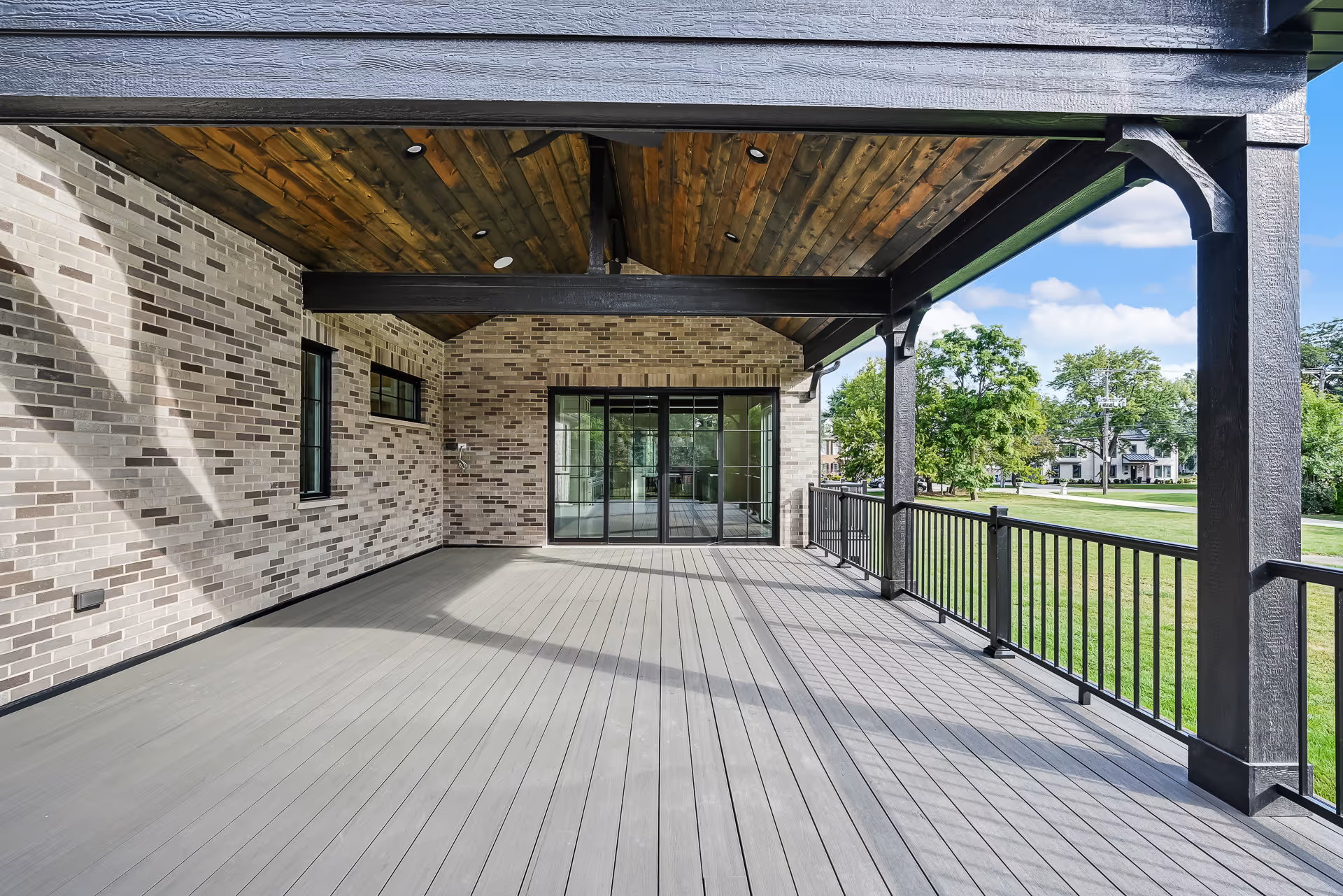 Spacious covered porch with light gray floor, brick walls, and wooden ceiling overlooking a green lawn.