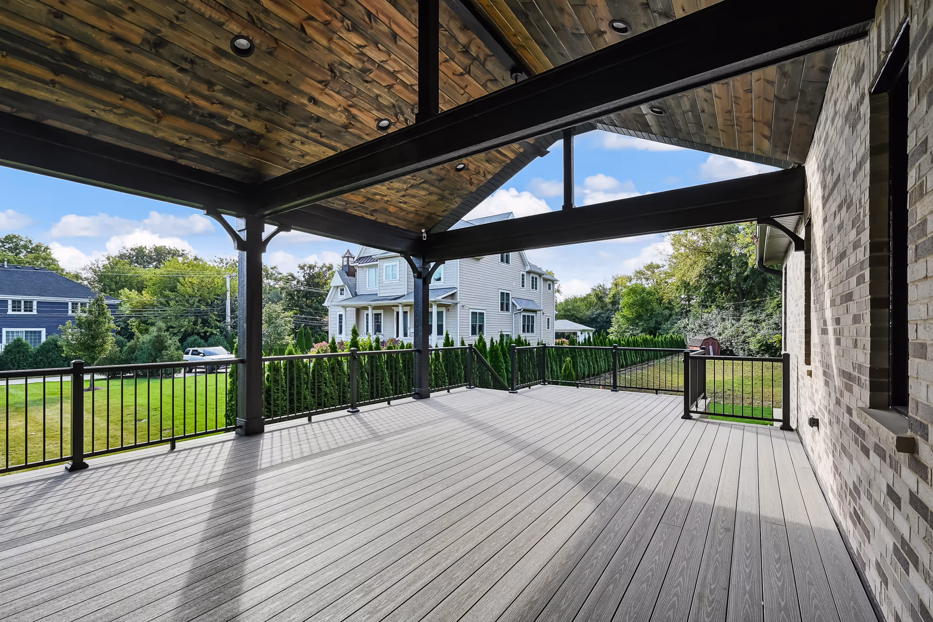 Spacious covered porch with wooden ceiling, gray deck flooring, black metal railing, and a view of neighboring houses and greenery.