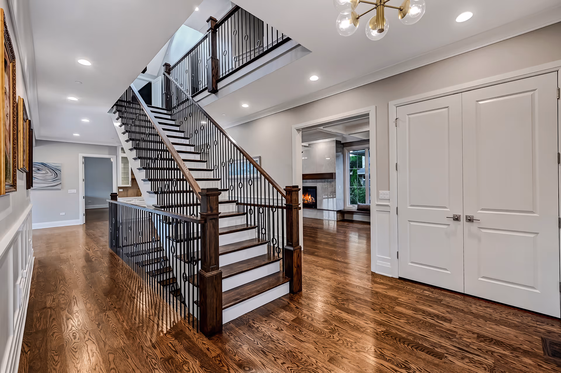 Interior of a modern home featuring wooden stairs with black iron railings, hardwood floors, a hallway with framed artwork, and a living room with a fireplace.