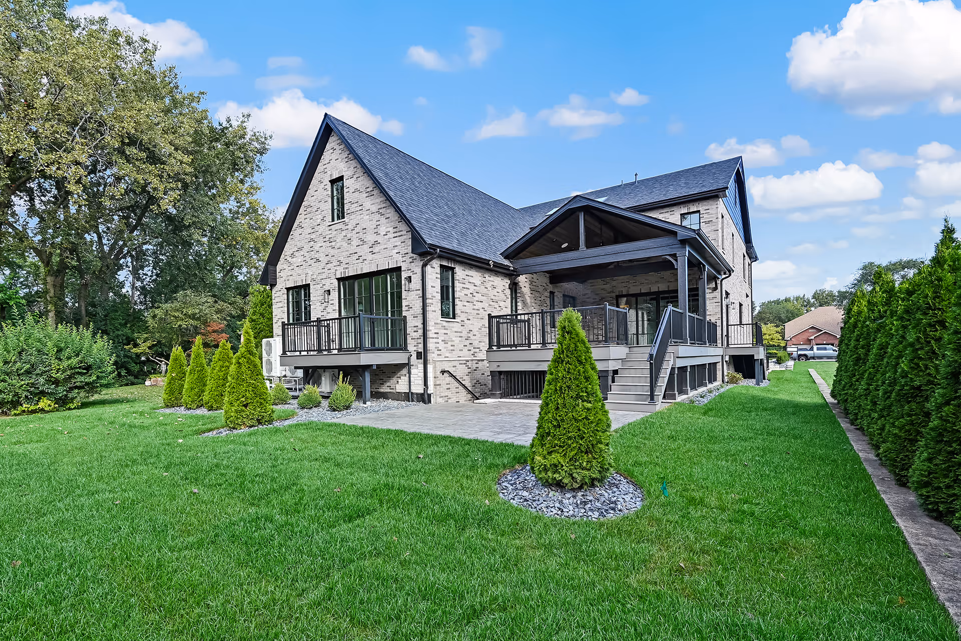 Modern light-colored brick house with dark roof, balconies, patio, and neatly manicured green lawn with small conical shrubs.