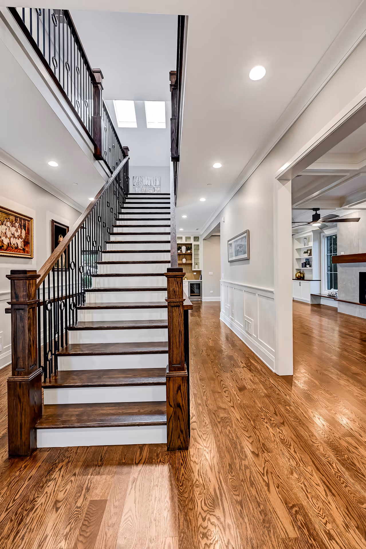 Interior view of a modern home featuring wooden stairs with white risers and black metal railings, hardwood floors, and a hallway leading to a living area with built-in shelves and a ceiling fan.