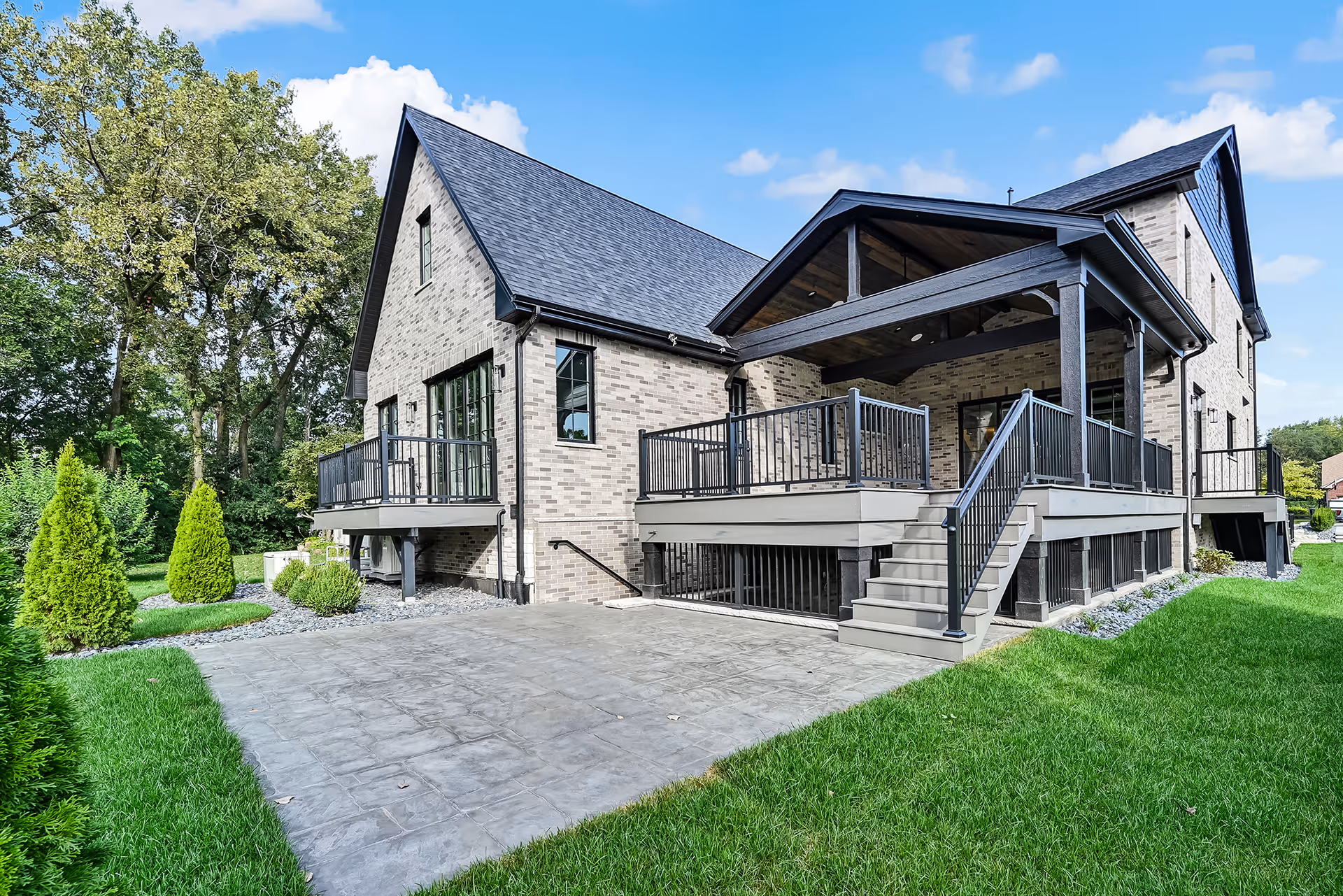 Modern two-story brick house with black roof, large deck with railing, and a concrete patio surrounded by green lawn and trees.