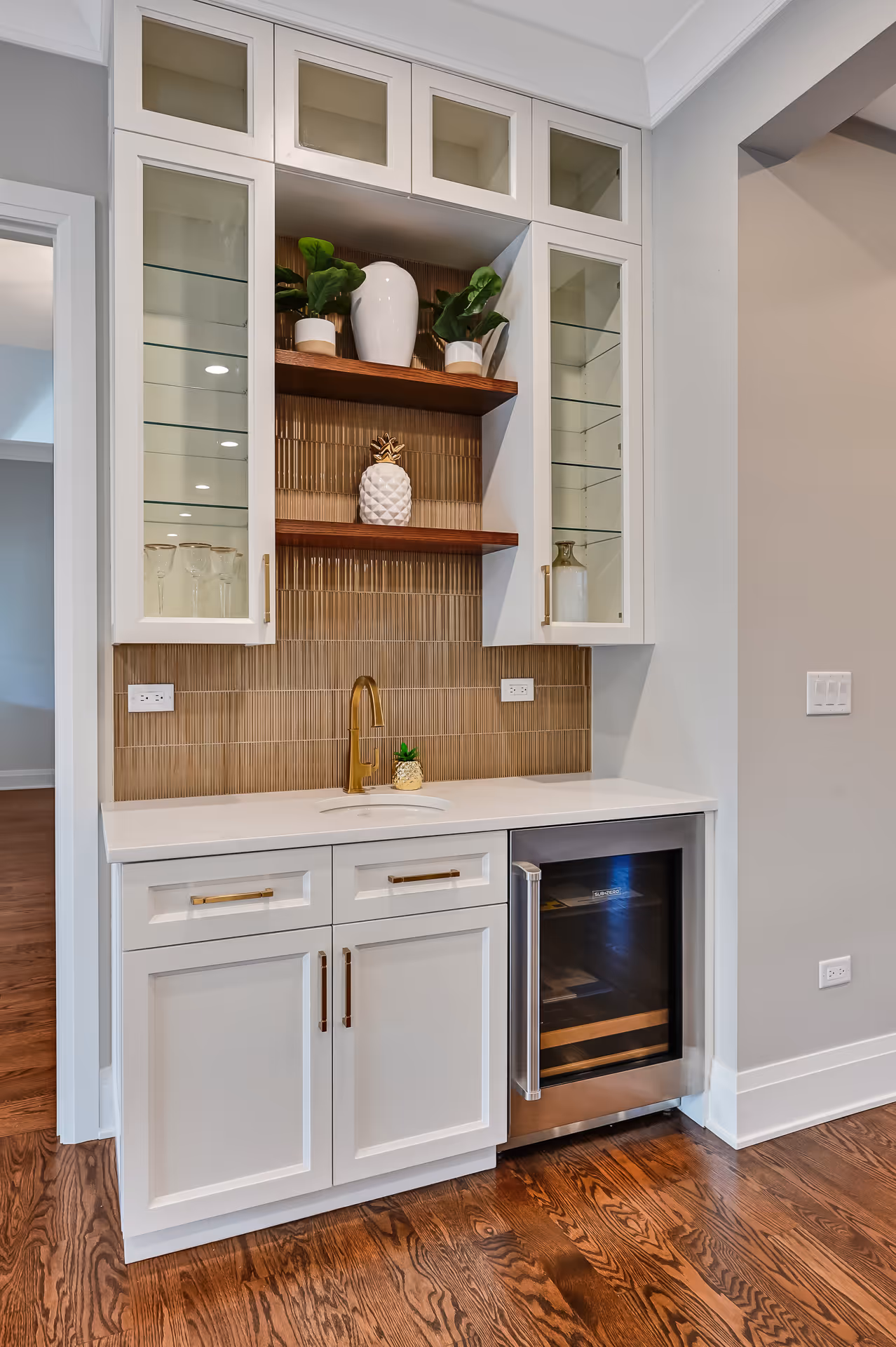 Modern white wet bar with gold faucet, wooden shelves displaying decorative vases and plants, glass-front cabinets, and a built-in stainless steel mini fridge on hardwood floor.