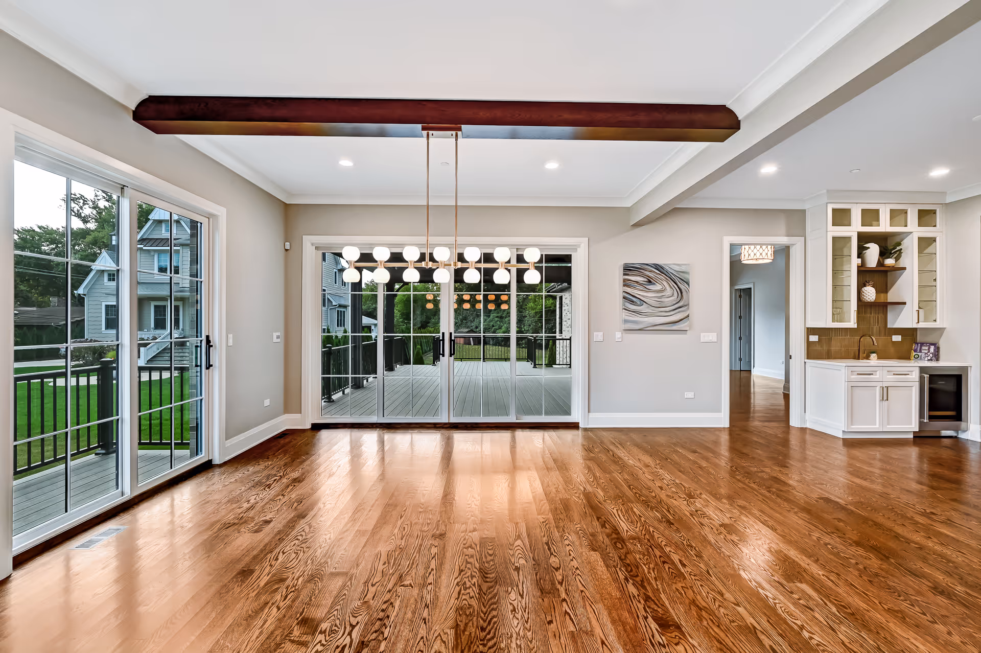 Spacious empty room with polished hardwood floors, large sliding glass doors opening to a deck, modern pendant light, and a small built-in cabinet area.