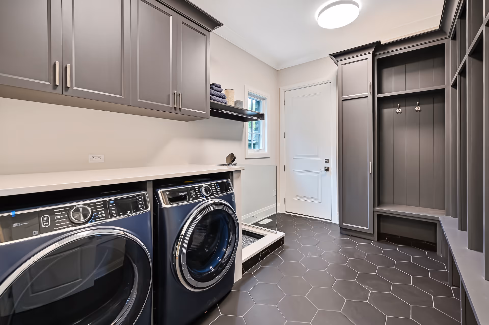 Modern laundry room with dark hexagonal floor tiles, front-loading washer and dryer, gray cabinetry, built-in bench with hooks, and a small window near a white door.