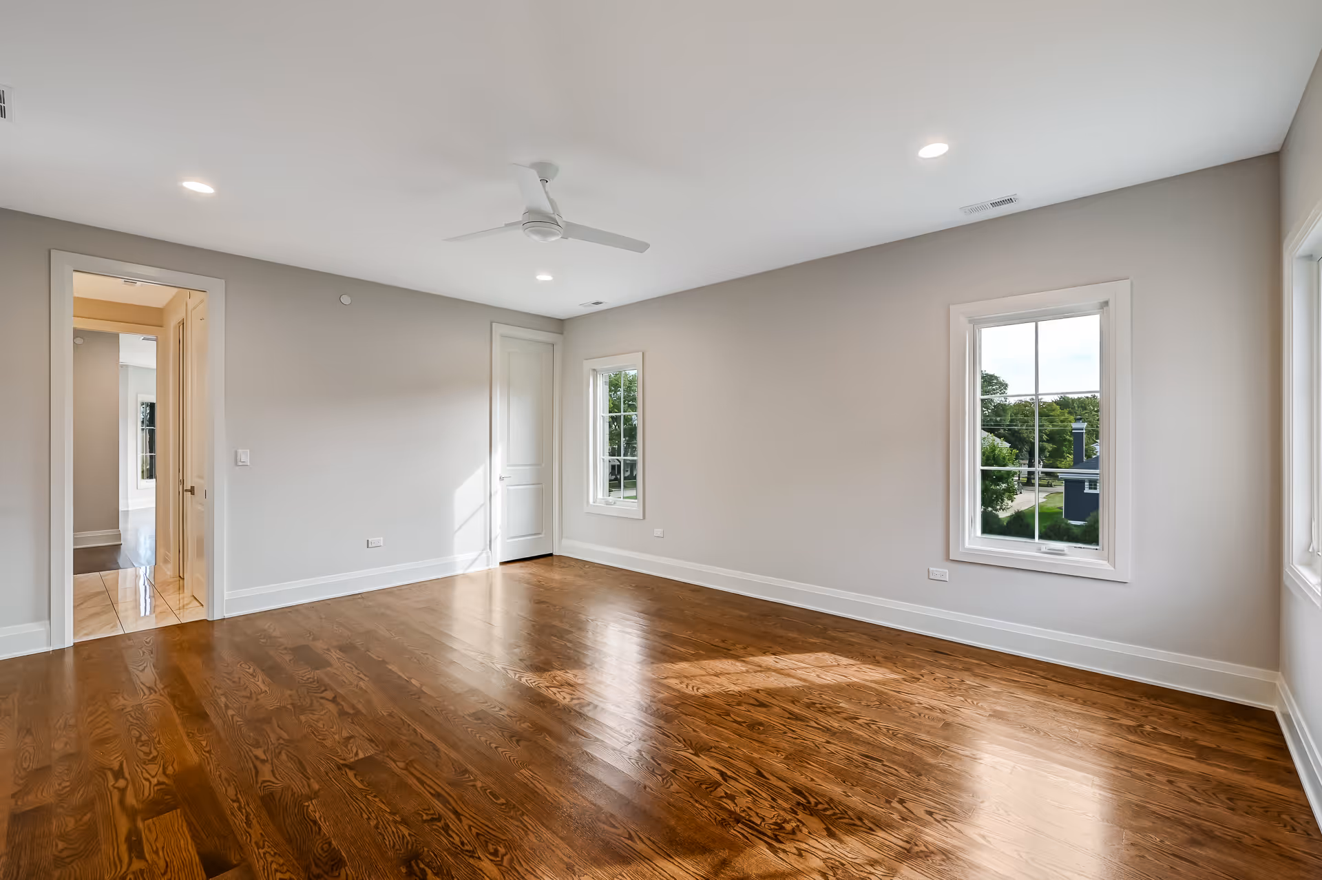 Empty room with polished hardwood floor, beige walls, ceiling fan, and two windows showing greenery outside.
