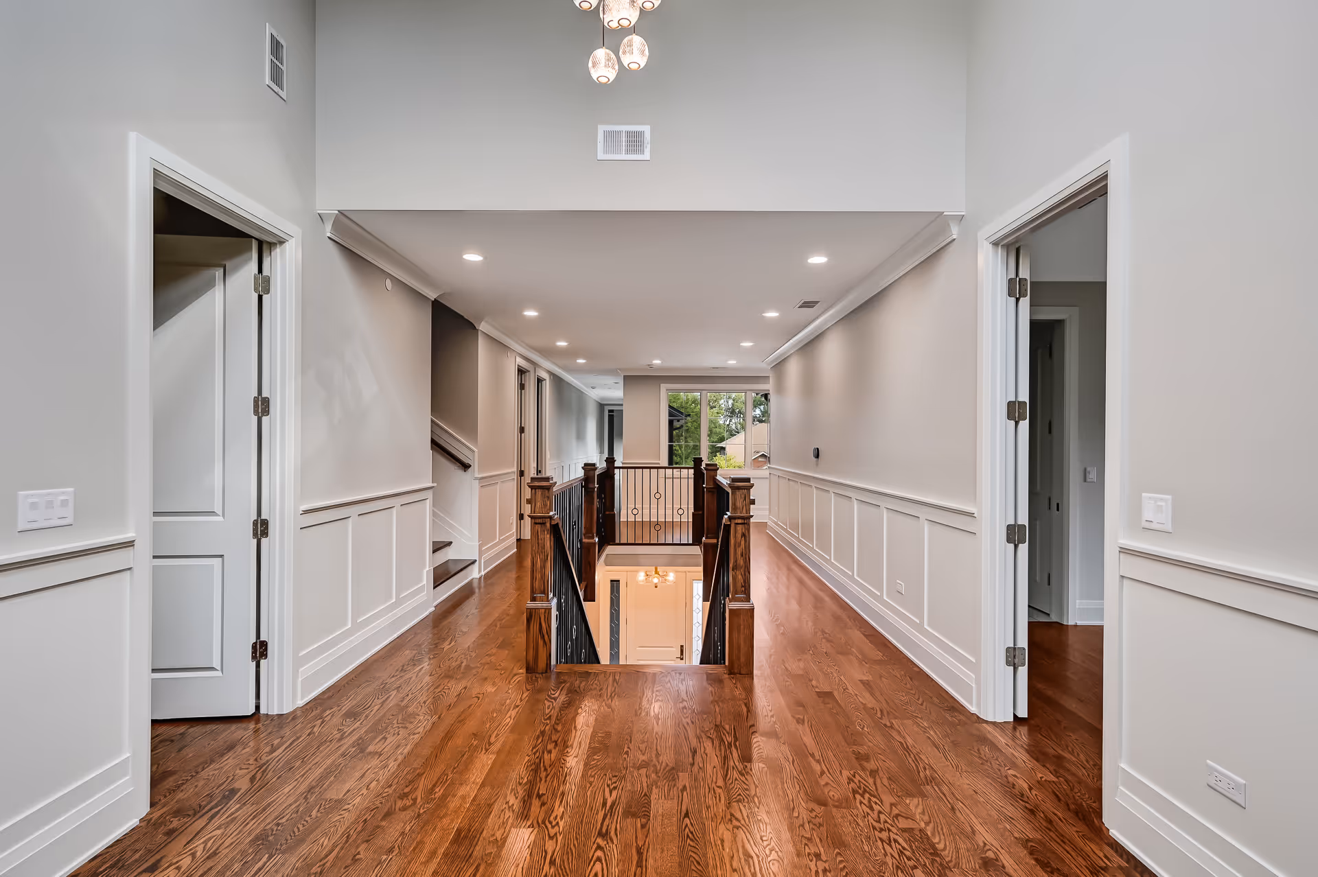 Spacious upper hallway with polished hardwood floors, white wainscoting, recessed ceiling lights, and wooden stair railings.