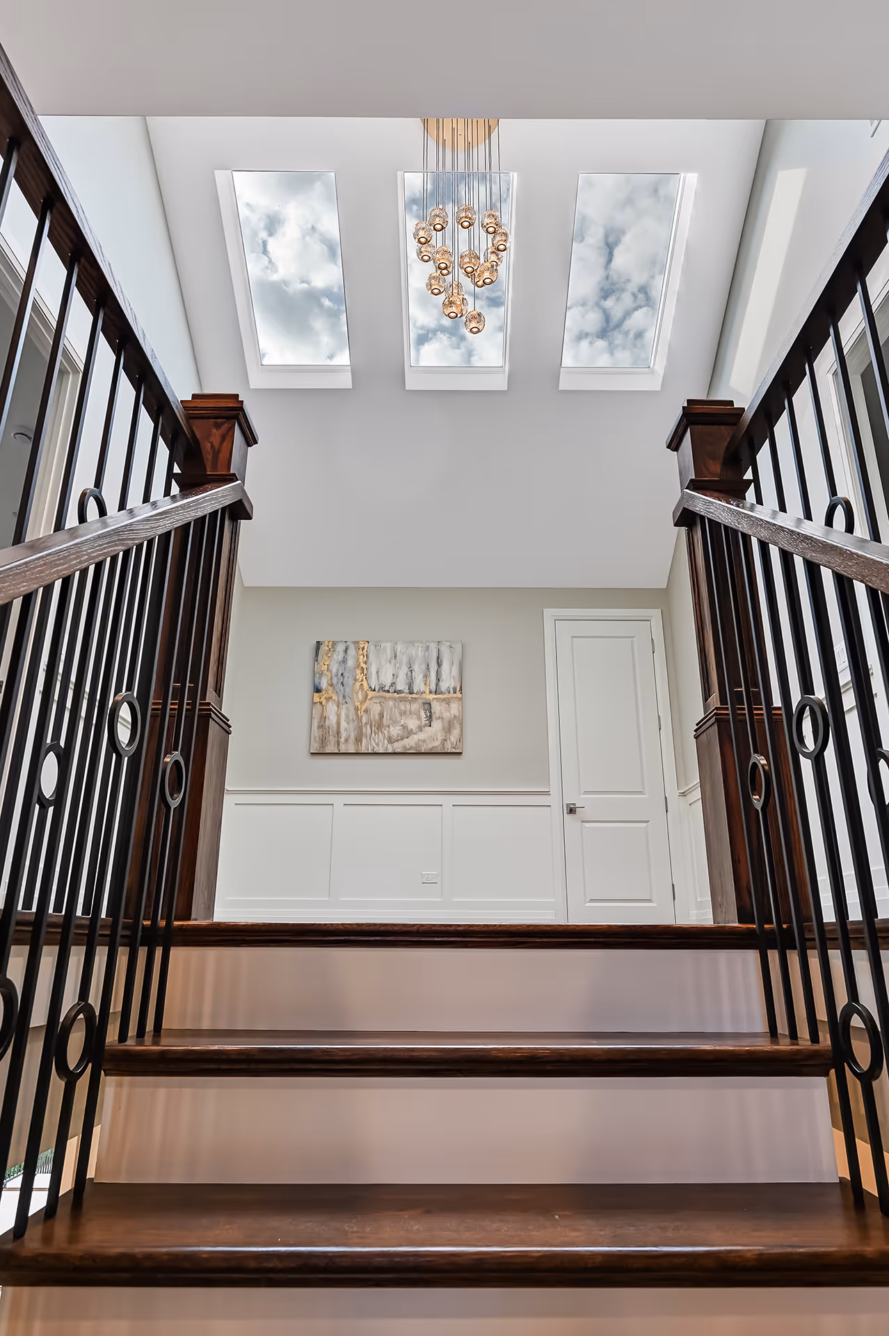 View from the base of wooden stairs with black iron railings leading to a landing with a white door, abstract wall art, and three skylights with a modern chandelier above.