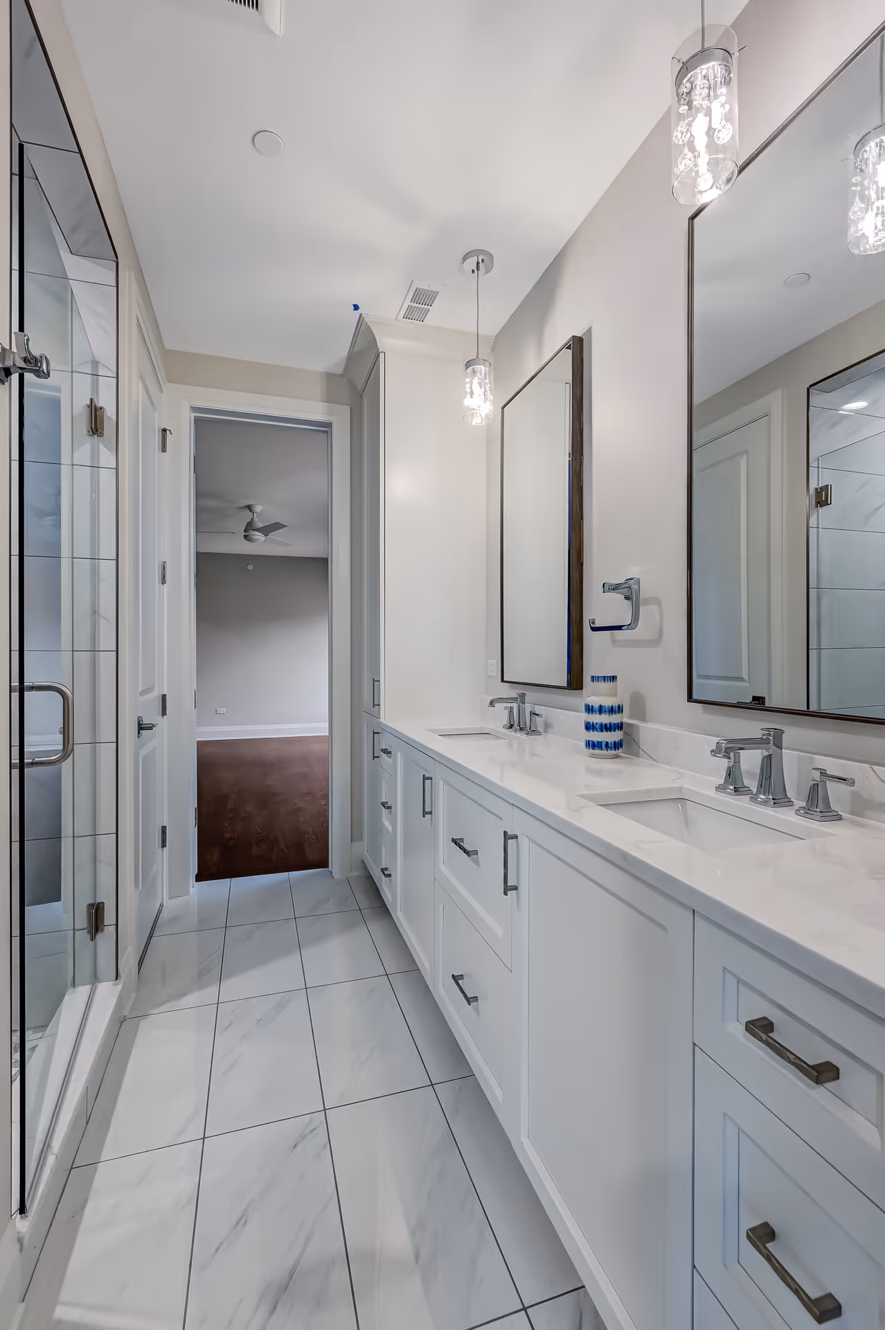 Modern bathroom with white double vanity, two mirrors, pendant lights, marble floor tiles, and glass shower door.