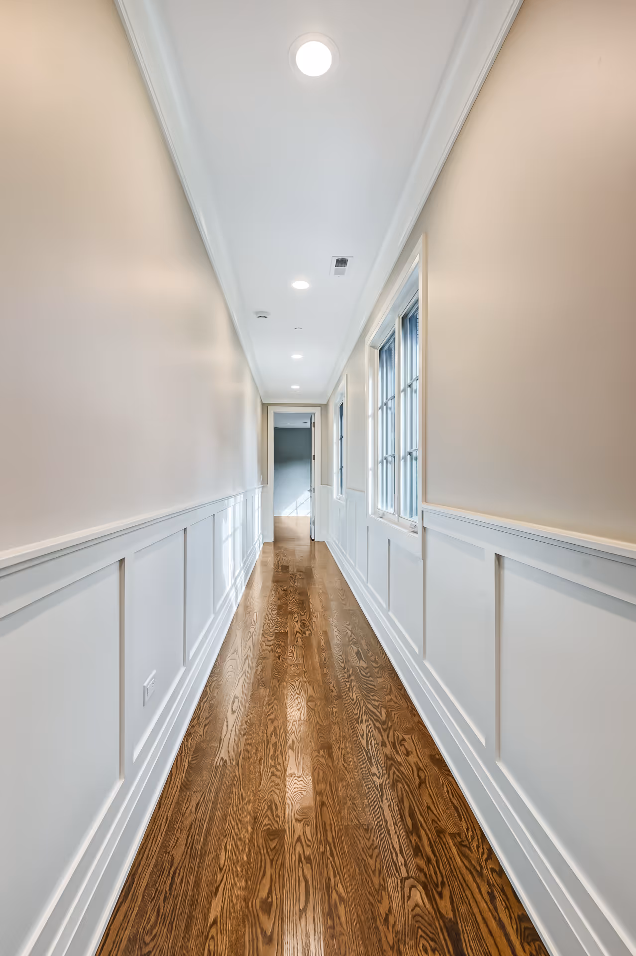 Long narrow hallway with white wainscoting, beige upper walls, hardwood floor, recessed ceiling lights, and windows on the right side.