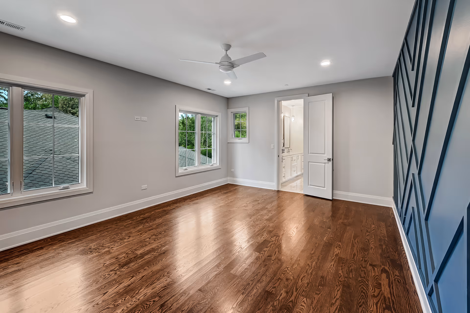 Empty room with polished hardwood floors, light gray walls, three windows, white ceiling fan, and an open door revealing a bathroom.