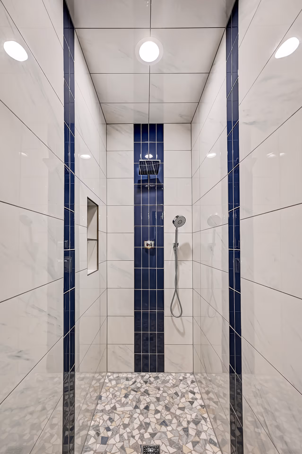Modern shower with white marble-effect tiles, vertical navy blue tile accents, a square overhead rain showerhead, handheld shower, and a mosaic tile floor.