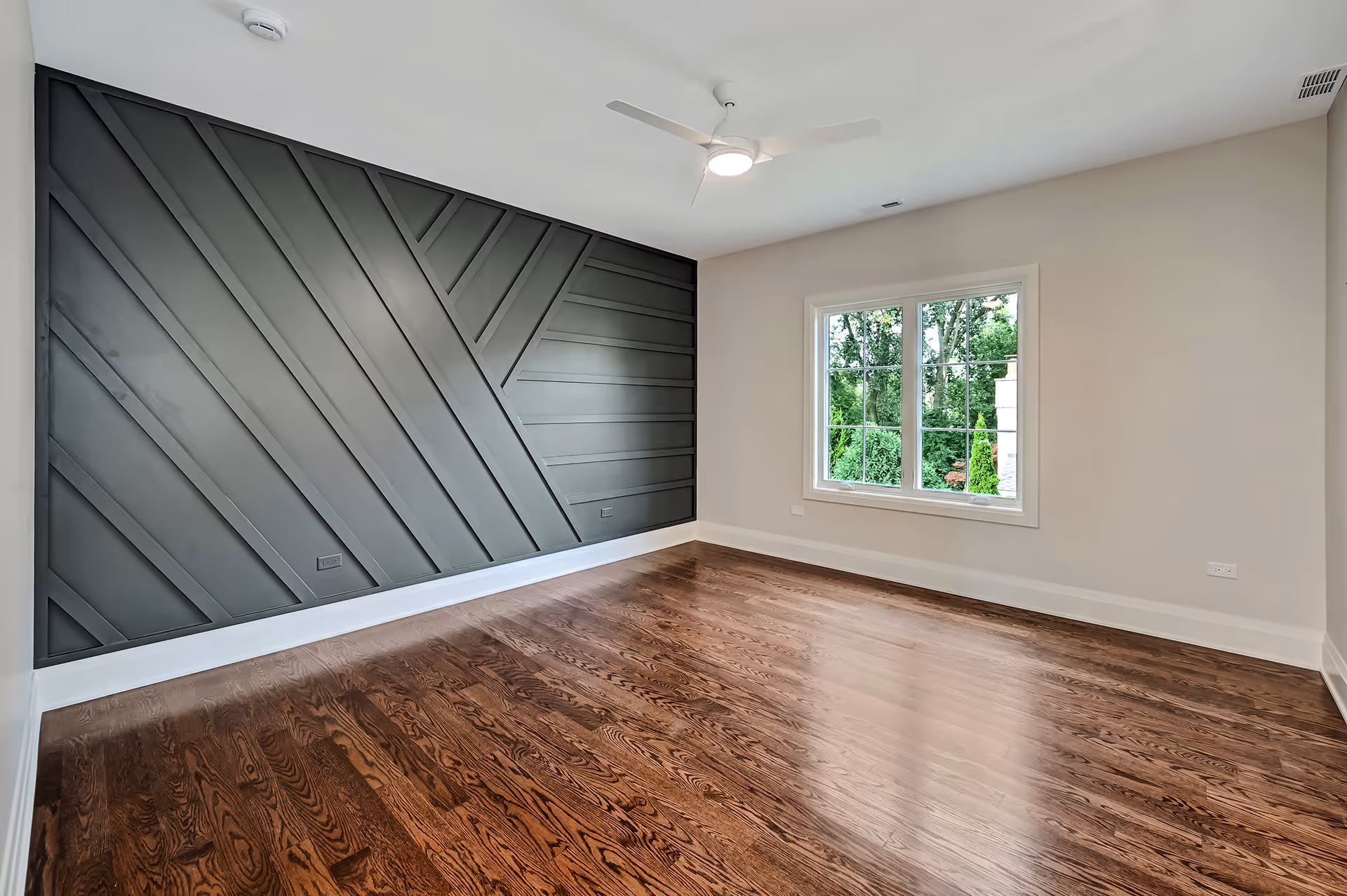 Empty room with dark wood floor, white walls, a large window, and a decorative dark gray paneled accent wall.