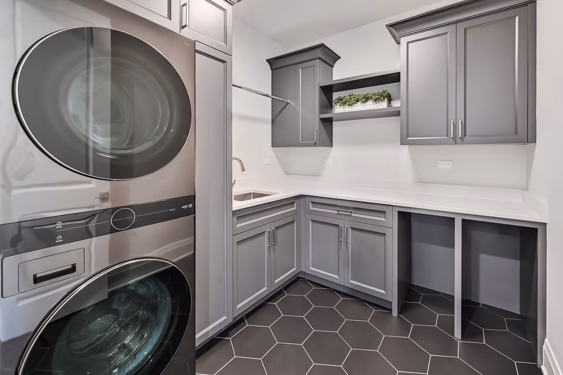 Modern laundry room with stacked front-loading washer and dryer, gray cabinetry, white countertop, and hexagonal black floor tiles.