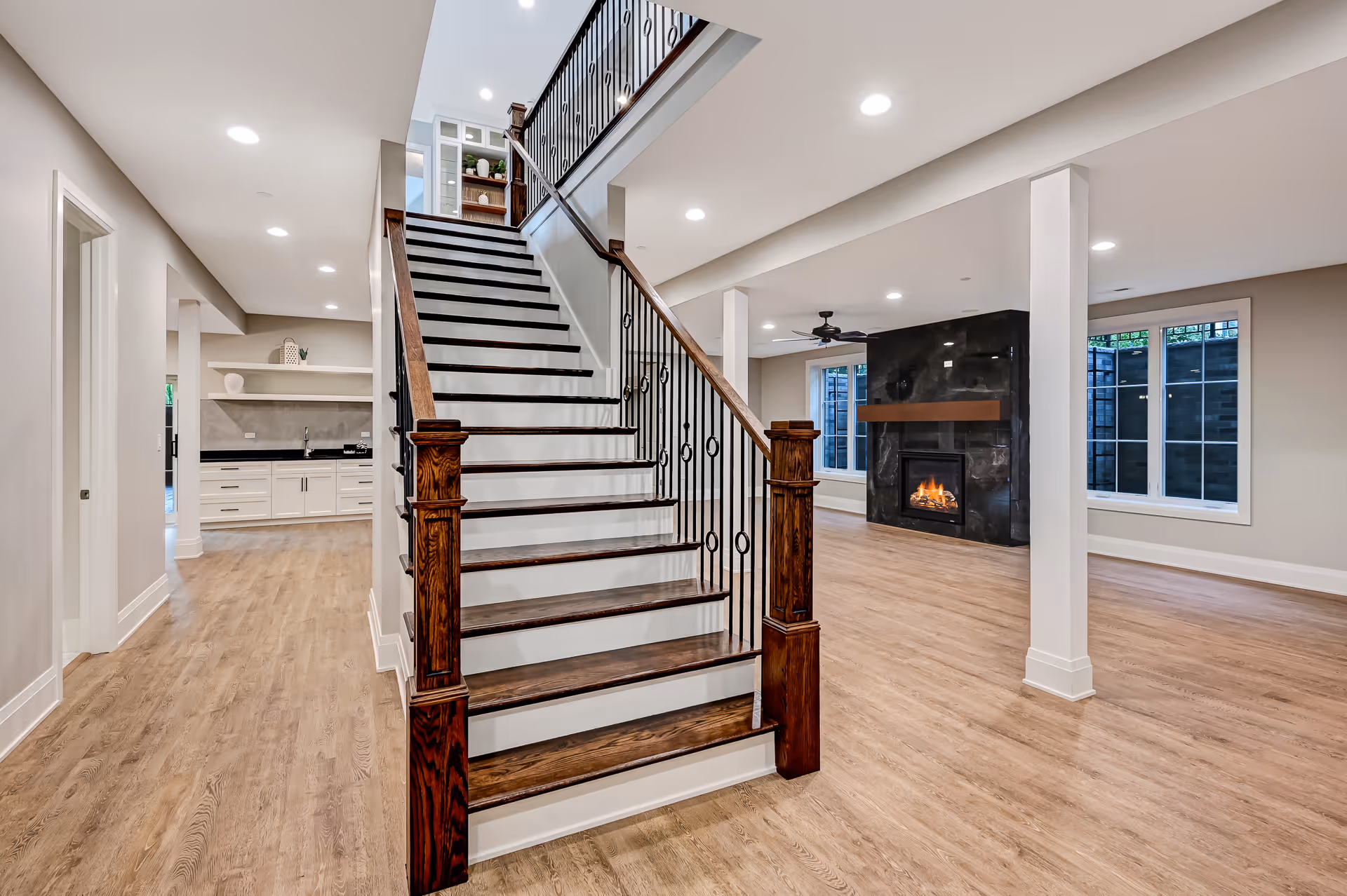 Modern open living space with wooden stairs, light wood flooring, a black stone fireplace, and recessed ceiling lights.