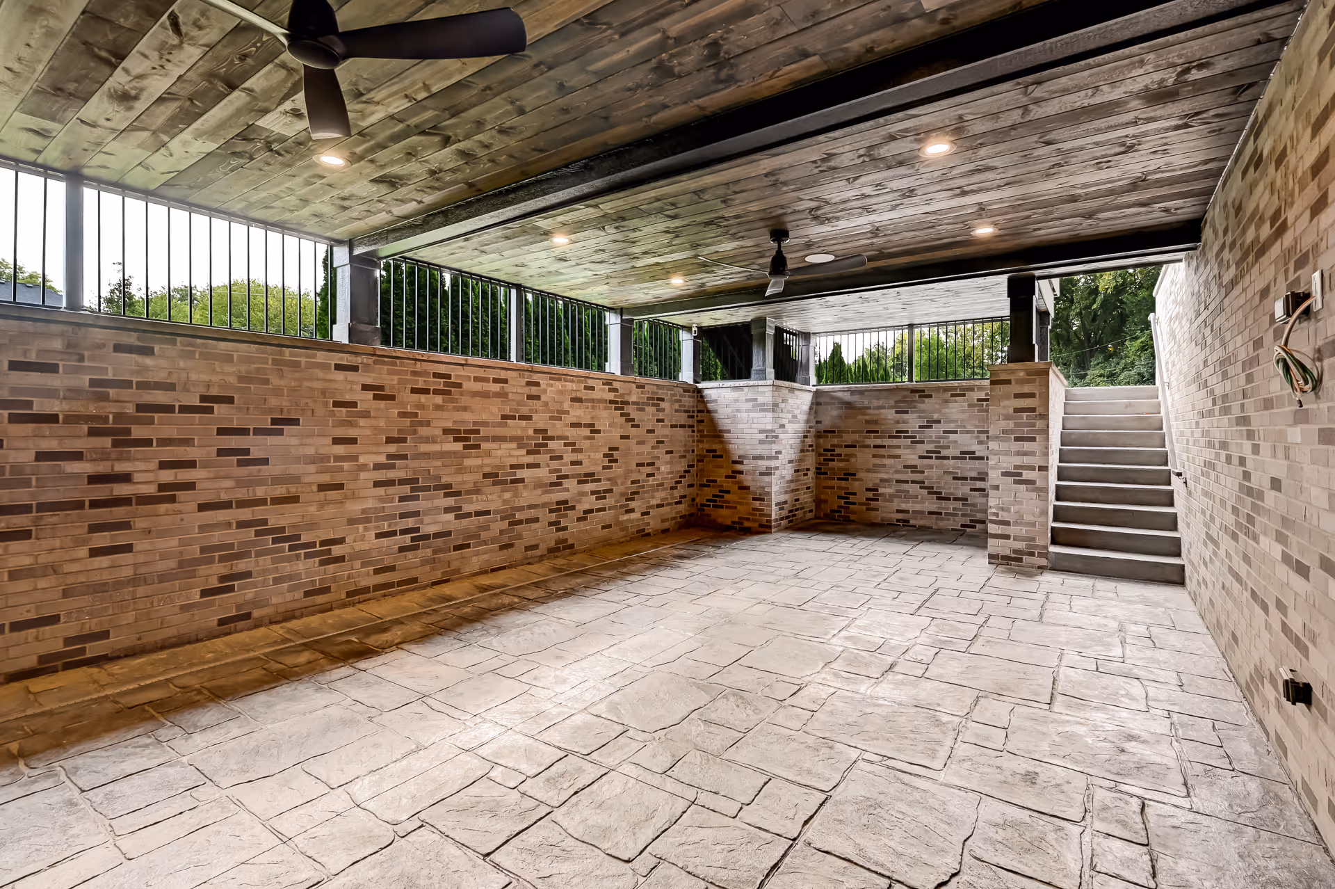 Covered outdoor patio area with brick walls, stamped concrete floor, ceiling fans, and stairs leading up to a garden.