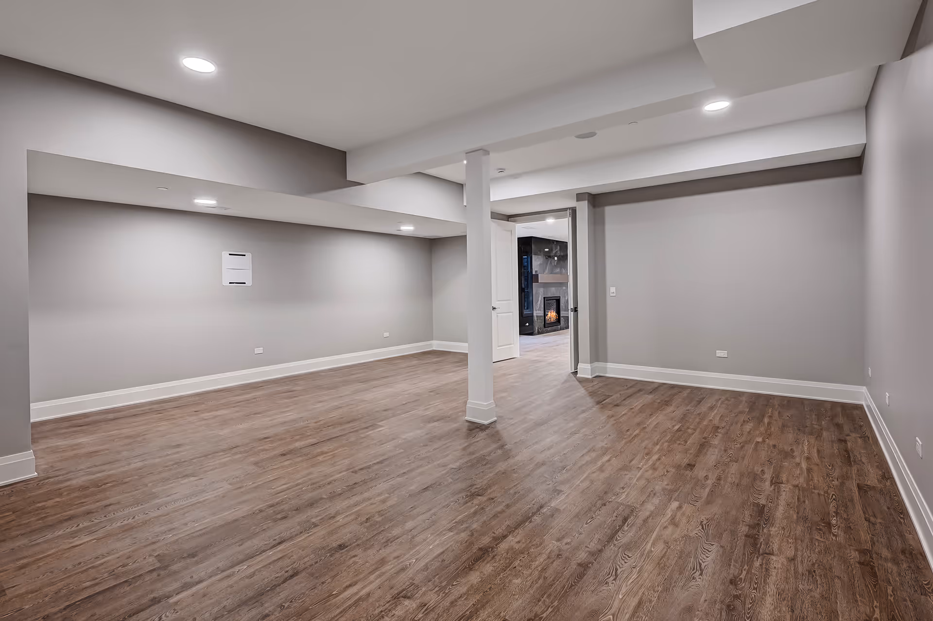 Empty basement room with gray walls, recessed ceiling lights, wood-look flooring, and a view through an open door showing a fireplace in the next room.