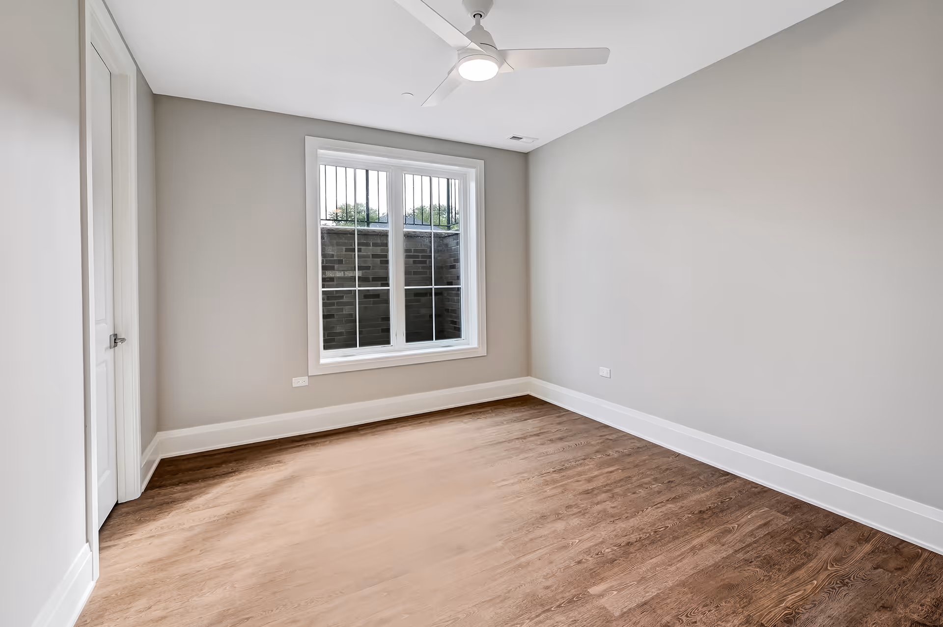 Empty room with gray walls, hardwood floor, a white ceiling fan, and a window showing a brick exterior wall.