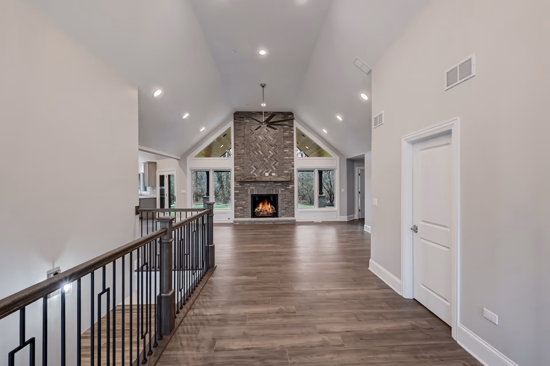 Spacious modern interior with vaulted ceiling, wood flooring, metal railing, and brick fireplace with fire burning.