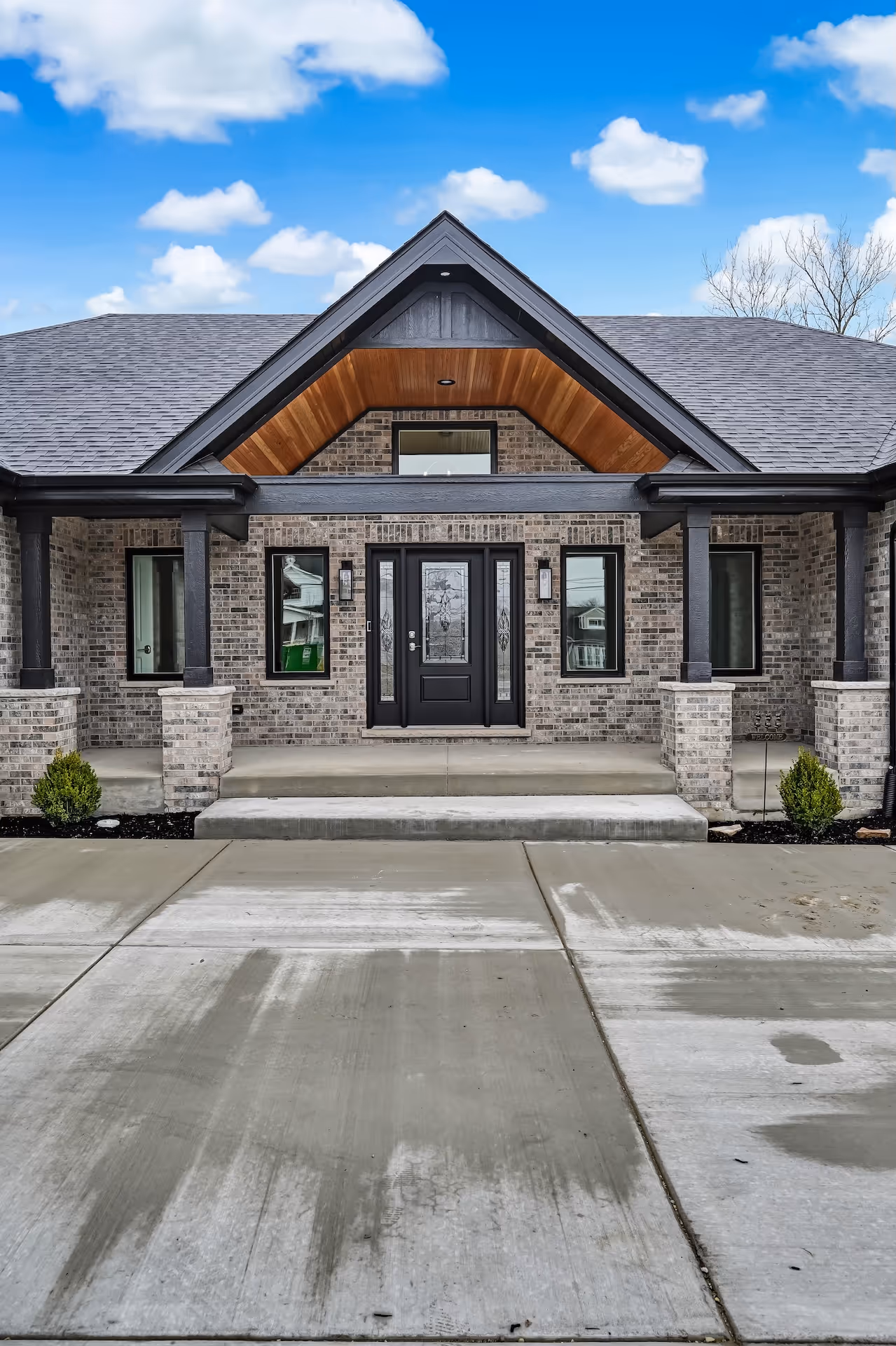 Front view of a brick house with a covered porch, black-framed windows, and a black door with decorative glass panels under a peaked roof with wood paneling.