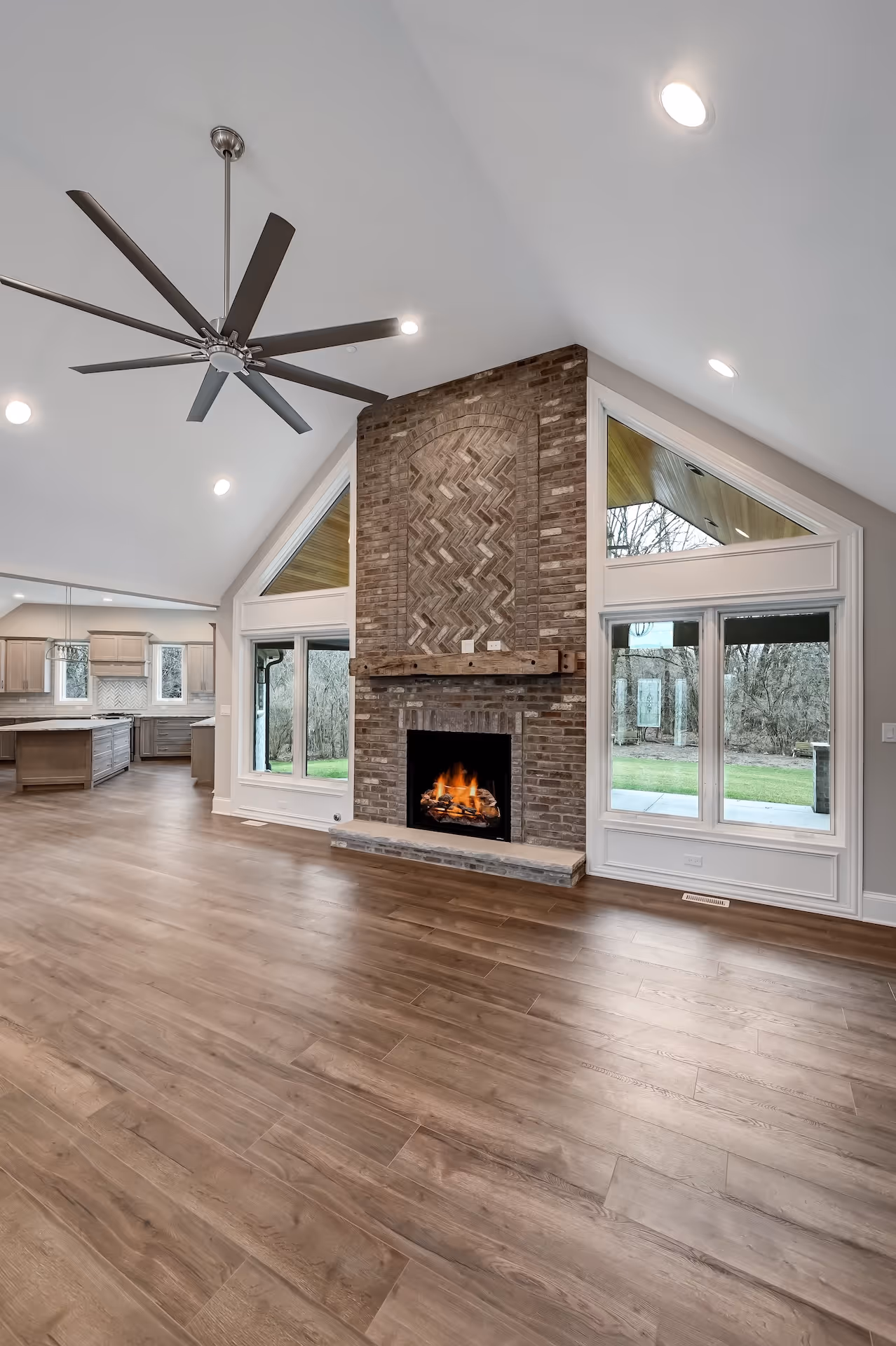 Spacious living room with a high vaulted ceiling, large ceiling fan, brick fireplace with fire, wood floor, and views of a kitchen and outdoors.