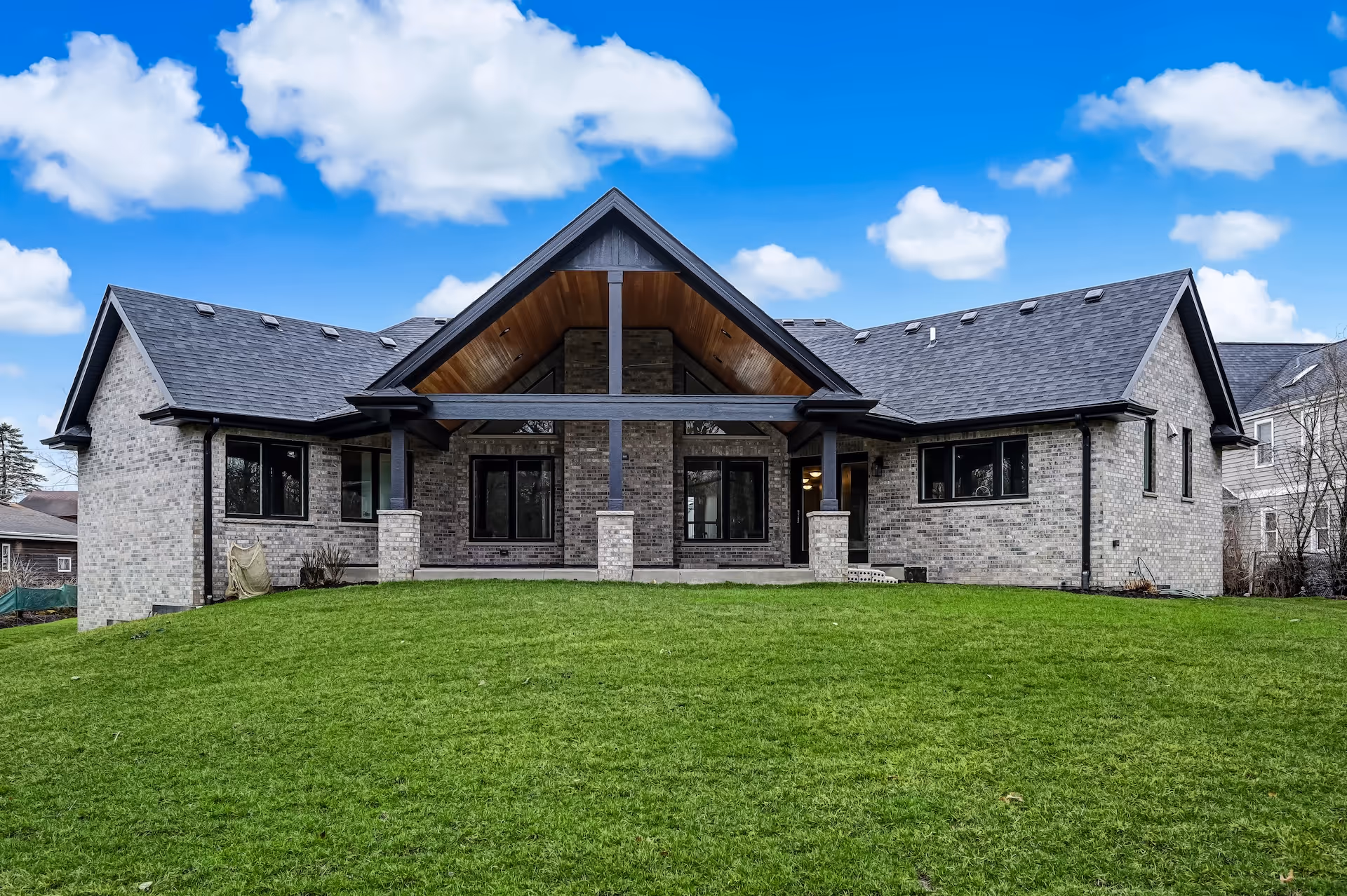 Modern single-story brick house with large covered front porch and expansive green lawn under a bright blue sky with scattered clouds.