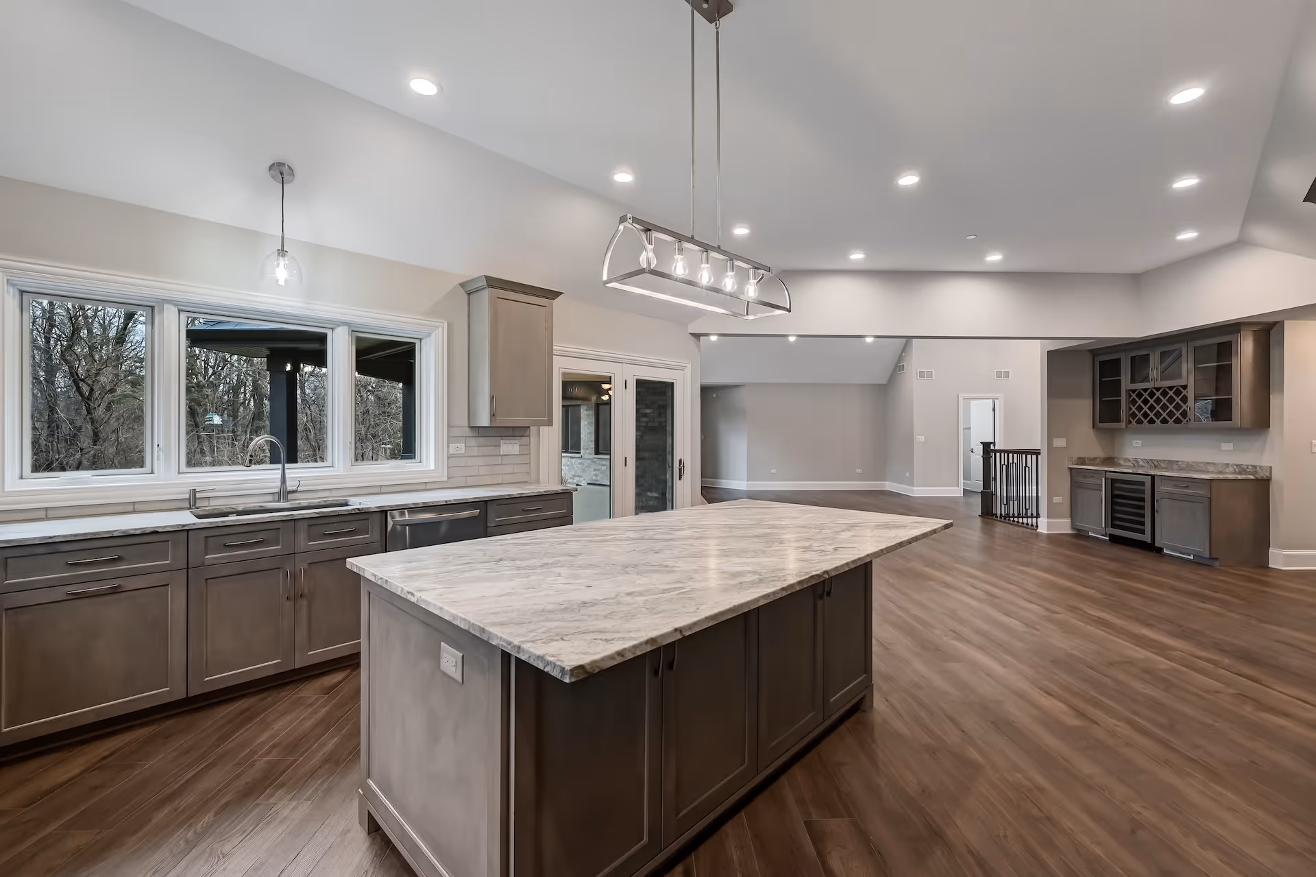 Spacious modern kitchen with large marble island, wood cabinets, hardwood floors, and large windows overlooking trees.