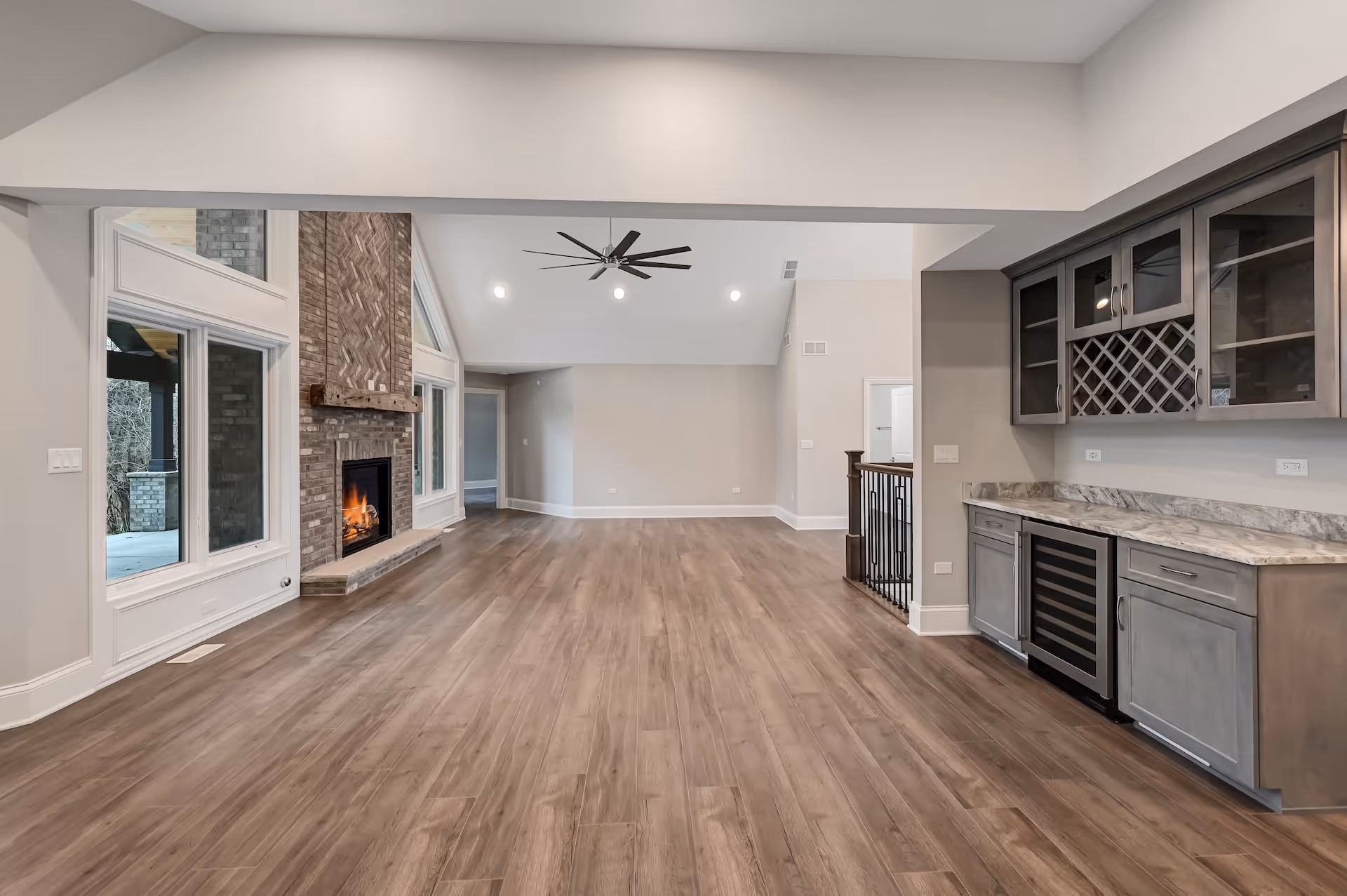 Spacious modern living room with wood flooring, a lit brick fireplace, large windows, ceiling fan, and a built-in wet bar with marble countertop.