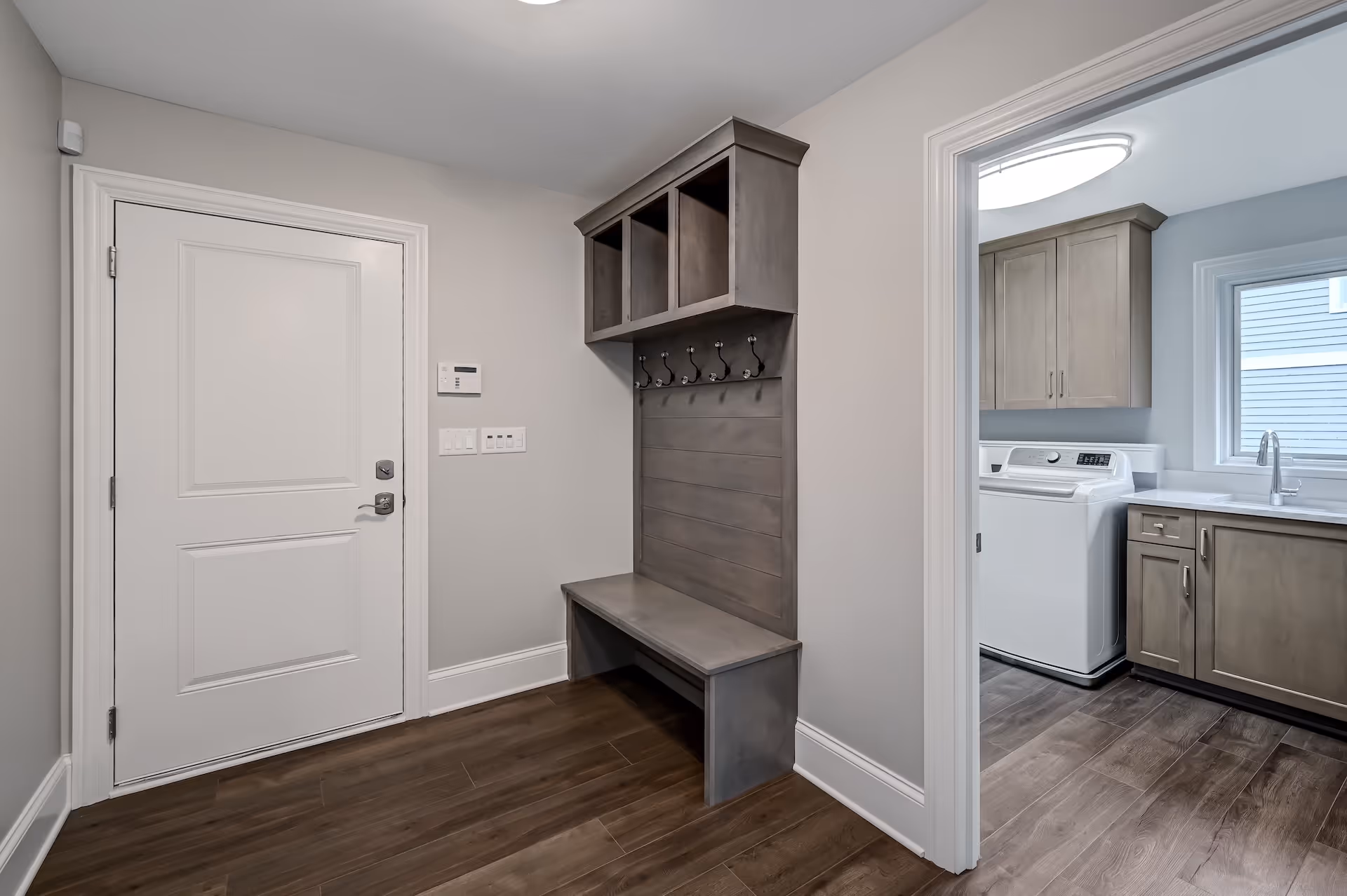 Entryway with a gray wooden bench and coat rack with cubby shelves, next to a white door and an adjacent laundry room with washer, cabinets, and a window.