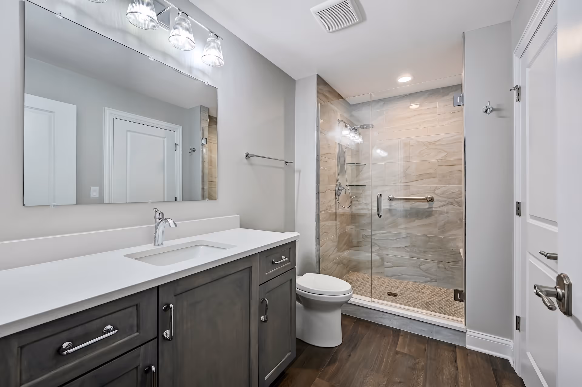 Modern bathroom with a large mirror, gray cabinets, white countertop, toilet, and glass-enclosed shower with beige tiles.