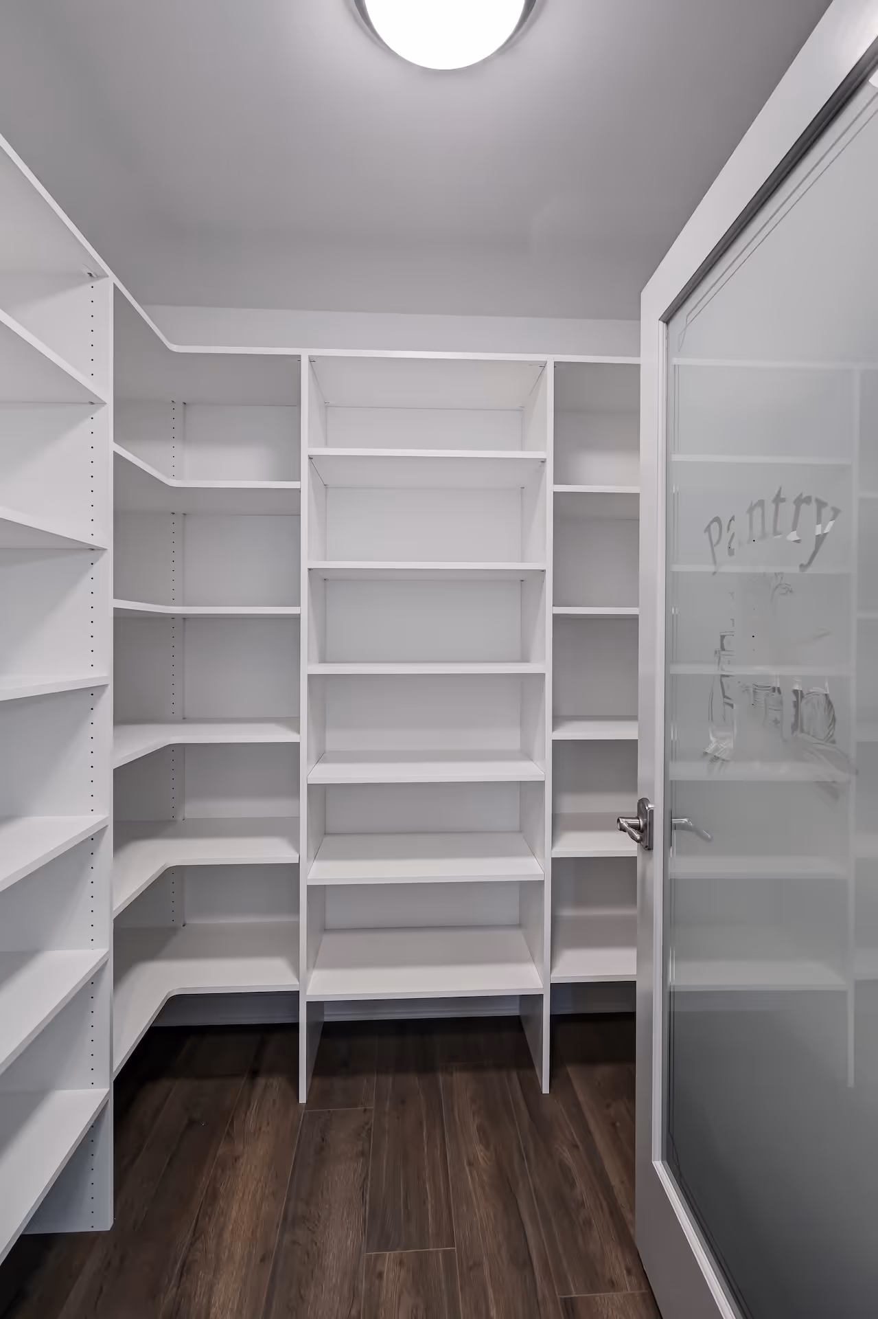 Empty white pantry shelves and wooden floor with a partially open frosted glass door labeled 'Pantry'.