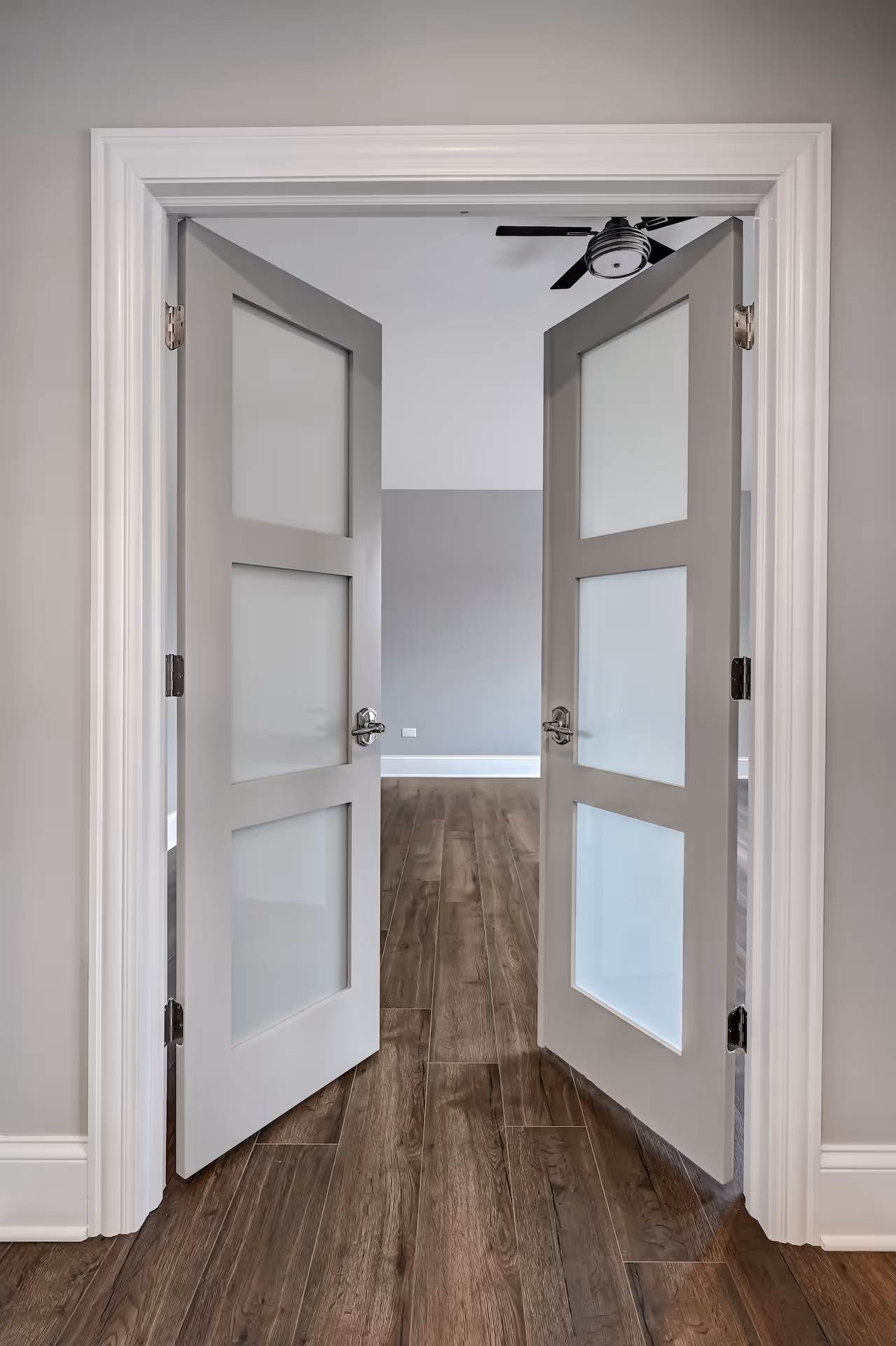 Open double doors with frosted glass panels opening to a room with gray walls, a ceiling fan, and wood flooring.