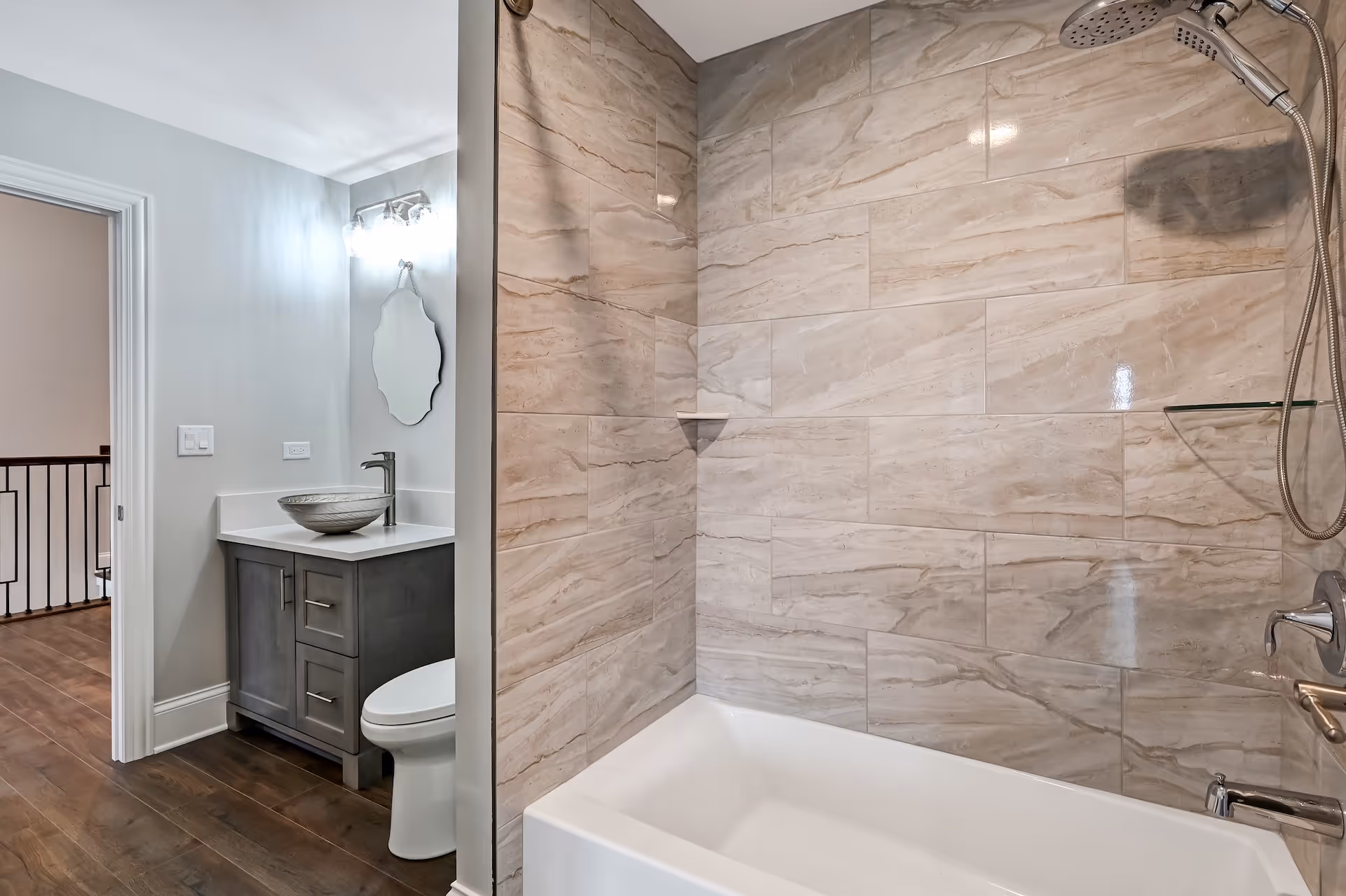 Modern bathroom with a white bathtub, beige marble wall tiles, gray vanity with vessel sink, and a round decorative mirror.