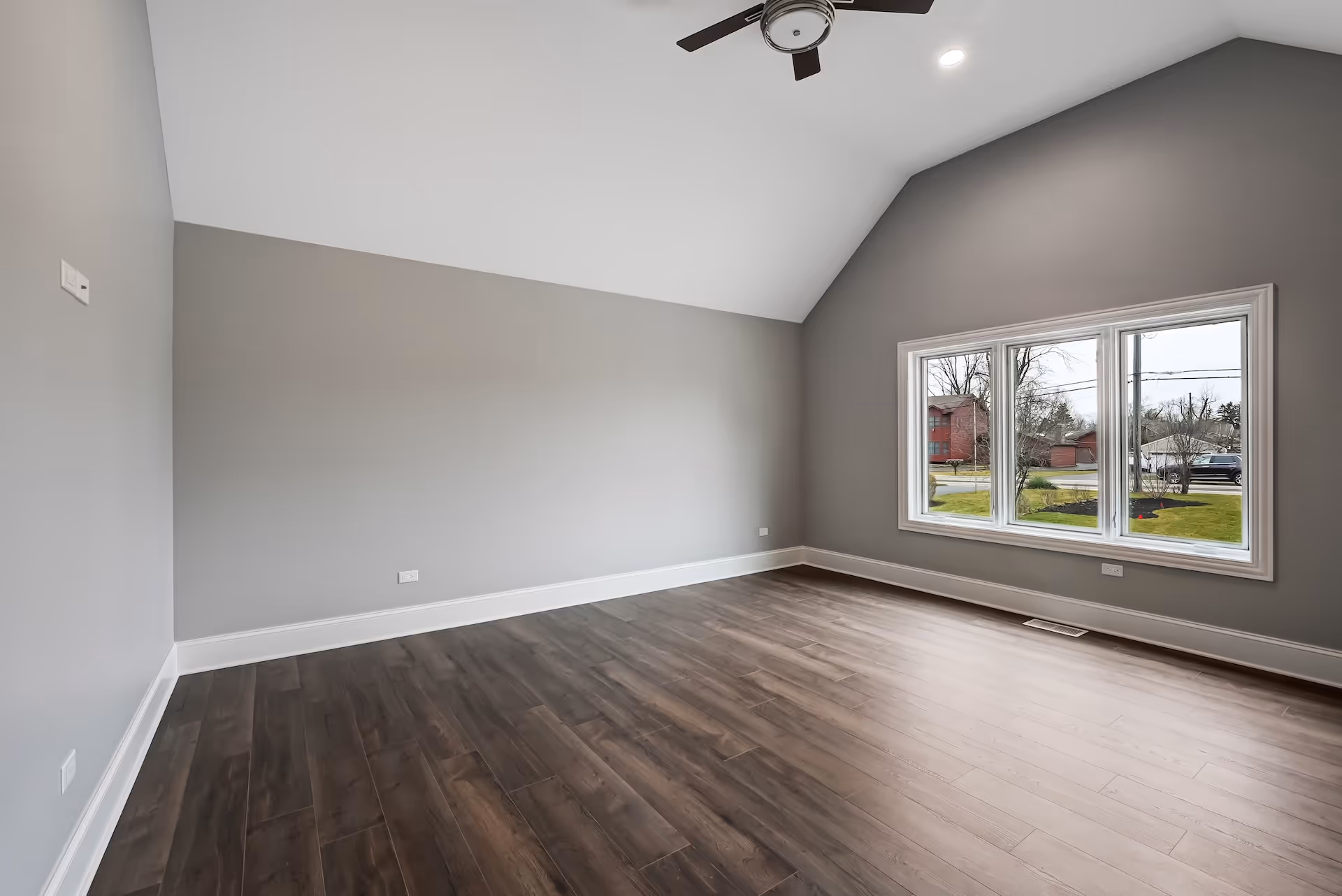 Empty room with vaulted ceiling, gray walls, dark hardwood floor, ceiling fan, and large window showing outside greenery and houses.
