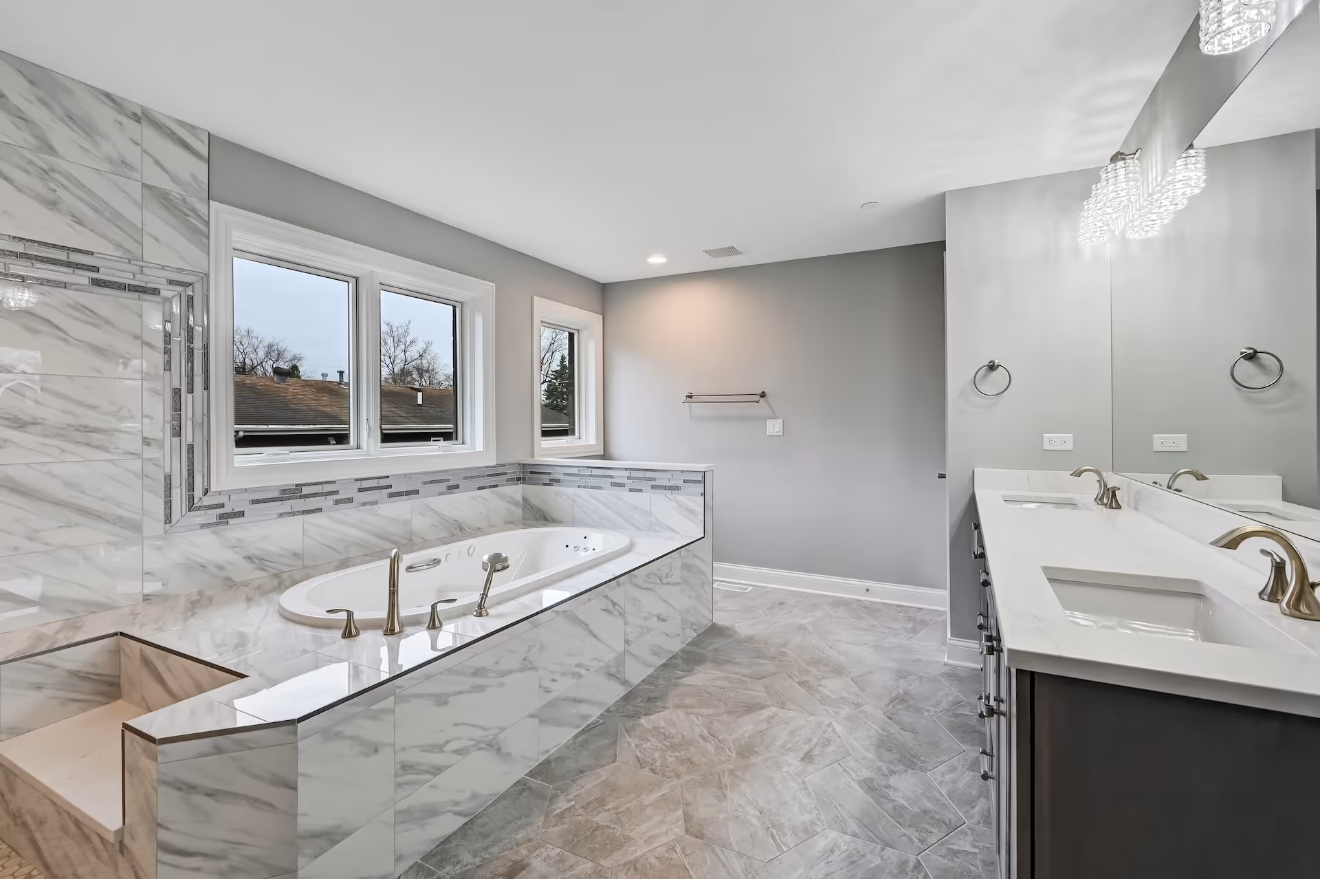 Modern bathroom featuring a white marble soaking tub with chrome fixtures, large mirror above double sinks, and gray tiled floor.