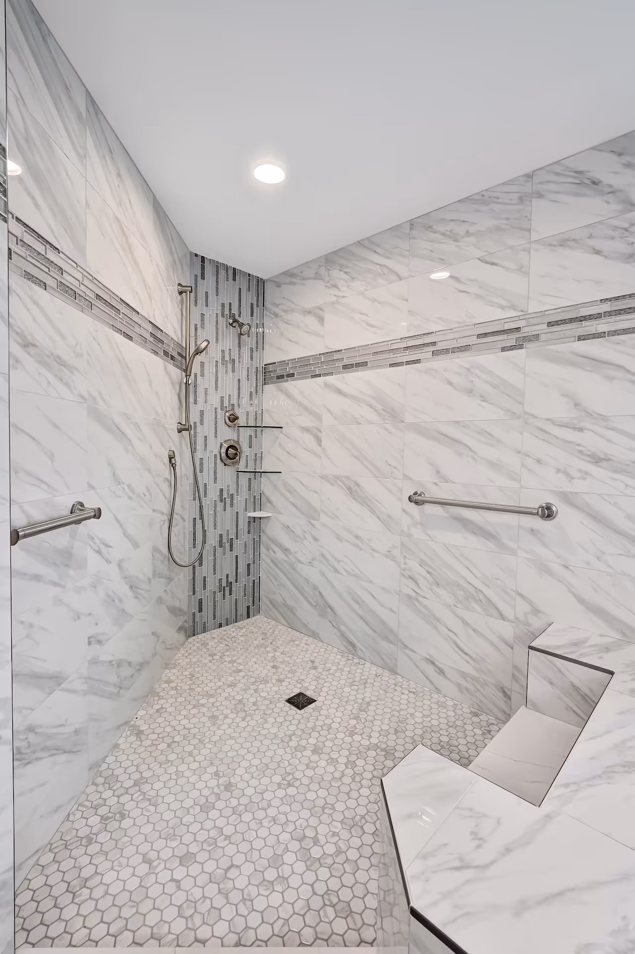 Modern walk-in shower with hexagonal tile floor, marble-patterned wall tiles, built-in shower bench, and stainless steel grab bars.