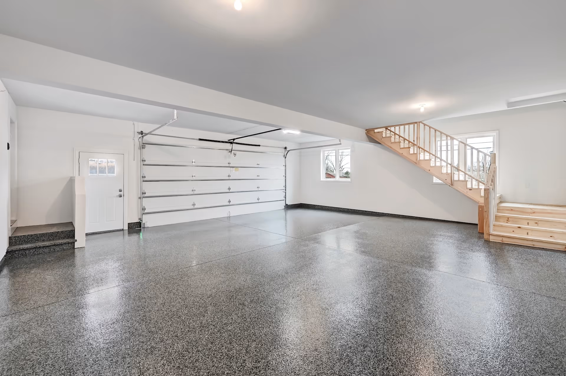 Clean, empty two-car garage with polished speckled gray floor, white walls, wooden staircase, and two small windows.
