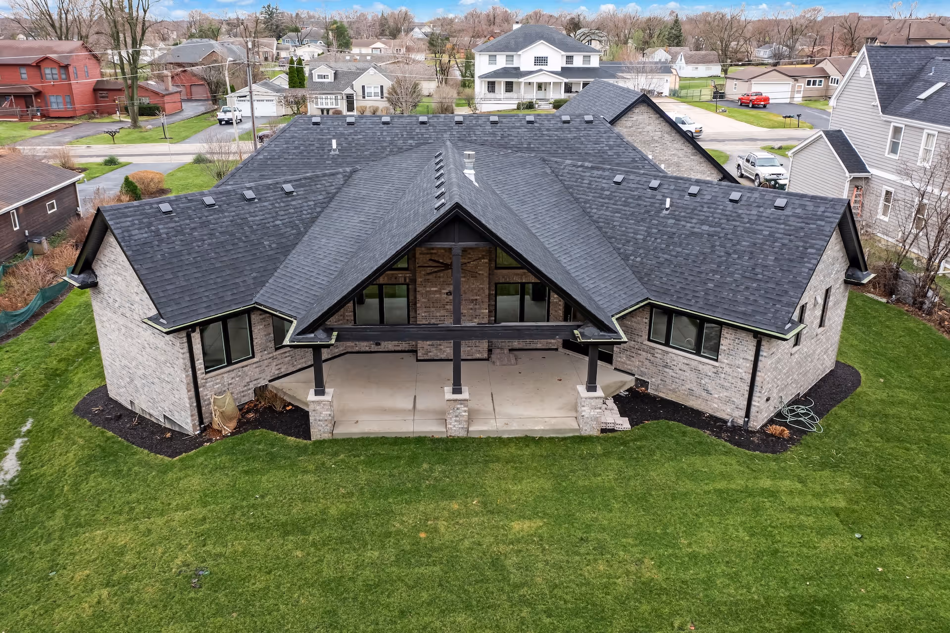 Aerial view of a modern brick house with dark gray shingle roof, large covered patio, and green lawn.