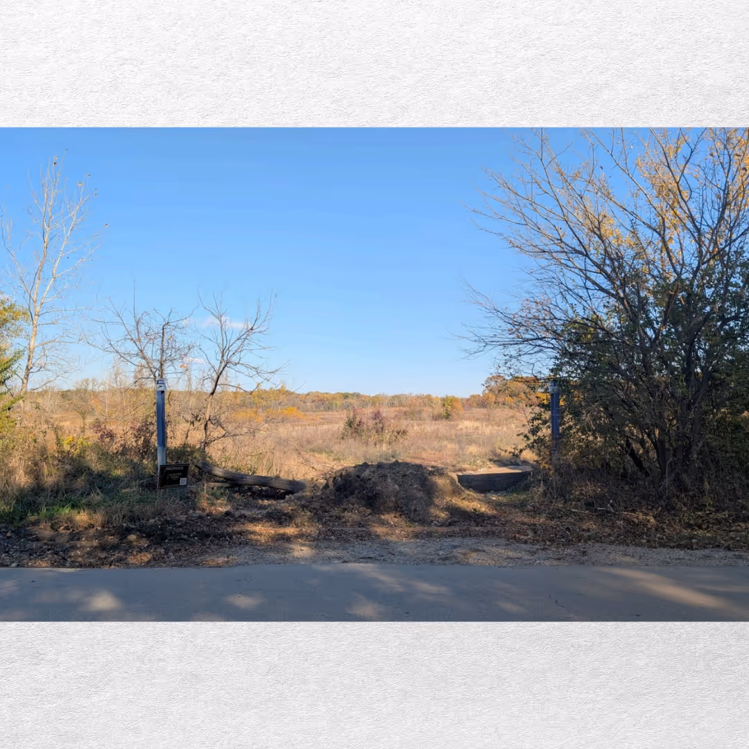 An undeveloped field with dry grass and scattered trees under a clear blue sky, seen beyond a dirt mound blocking a gravel path from a paved road.