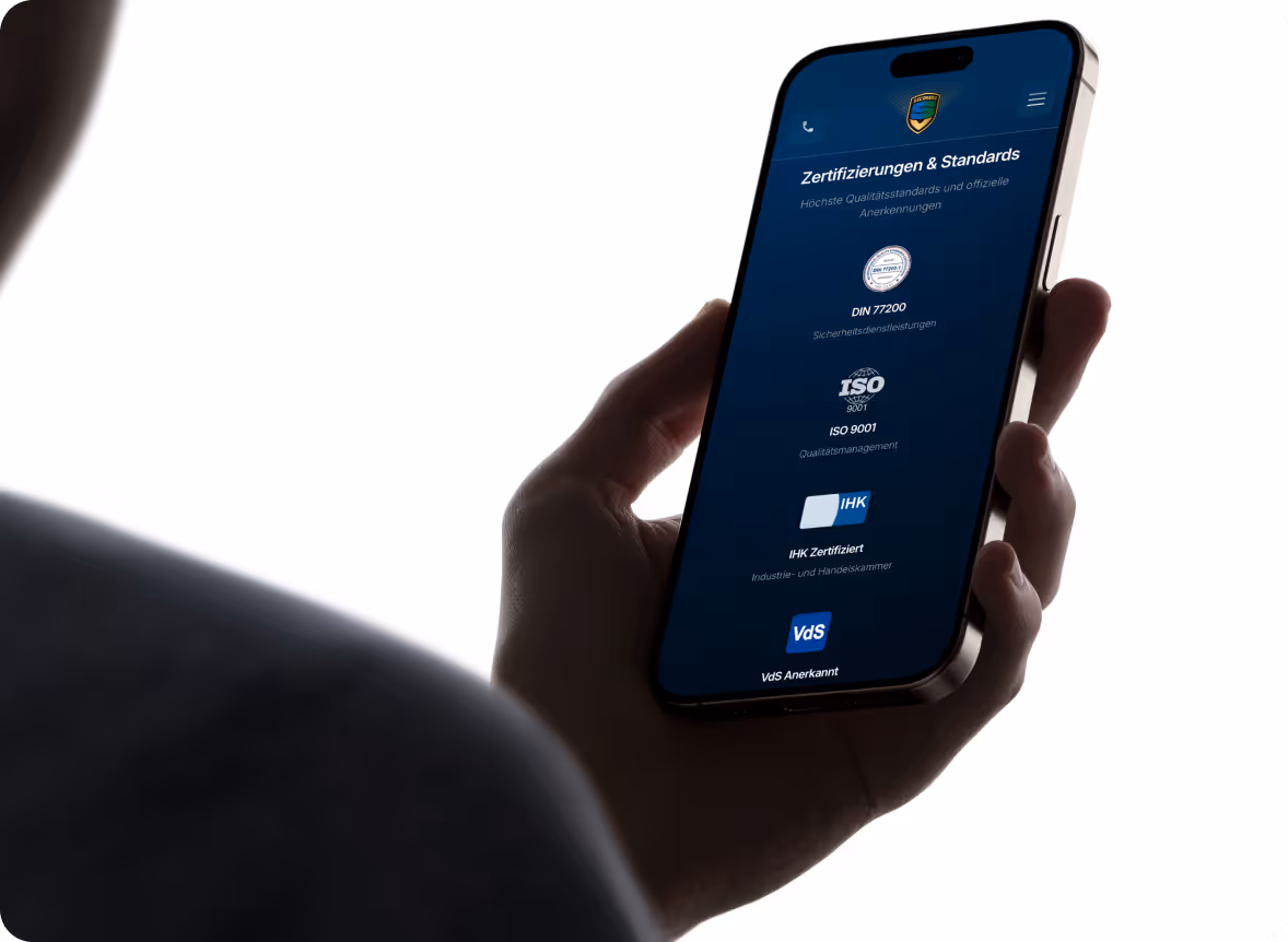 Hand hält ein Smartphone, das auf dem Bildschirm Zertifizierungen und Standards wie DIN 77200, ISO 9001, IHK Zertifiziert und VdS Anerkannt anzeigt.