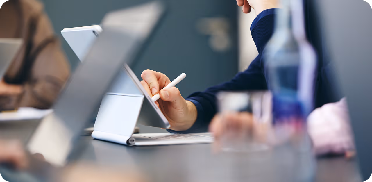 Person benutzt einen Stift, um auf einem Tablet mit Tastatur zu schreiben, während andere Personen ebenfalls digitale Geräte verwenden.