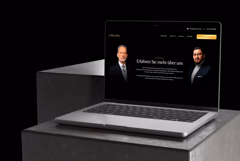 Aufgeklappter Laptop auf grauer Oberfläche mit der Webseite von GoldmanTax, die zwei Männer und Text 'Erfahren Sie mehr über uns' zeigt.