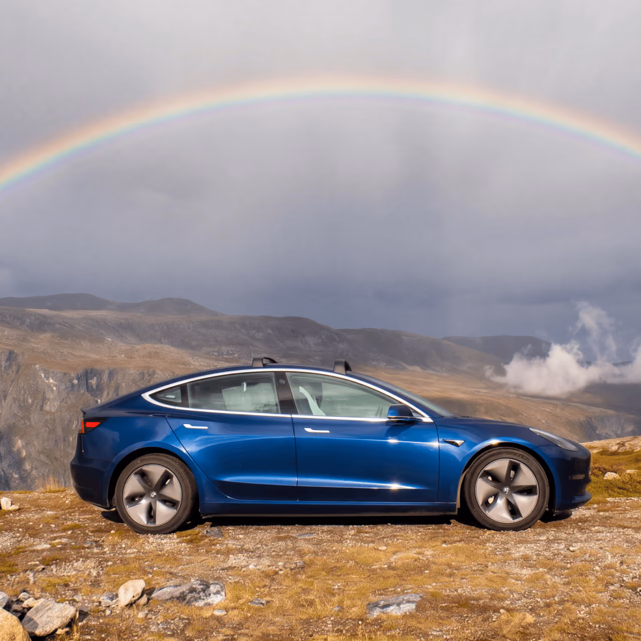 Blue Tesla Model 3 on a mountain meadow under a rainbow.